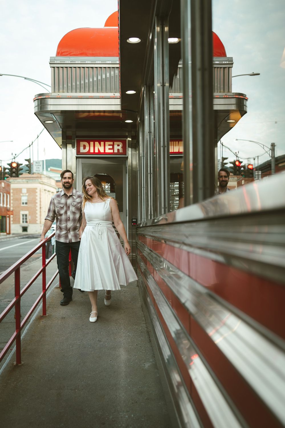 Couple walking outside a retro diner with red neon sign, woman in white dress, man in plaid shirt.
