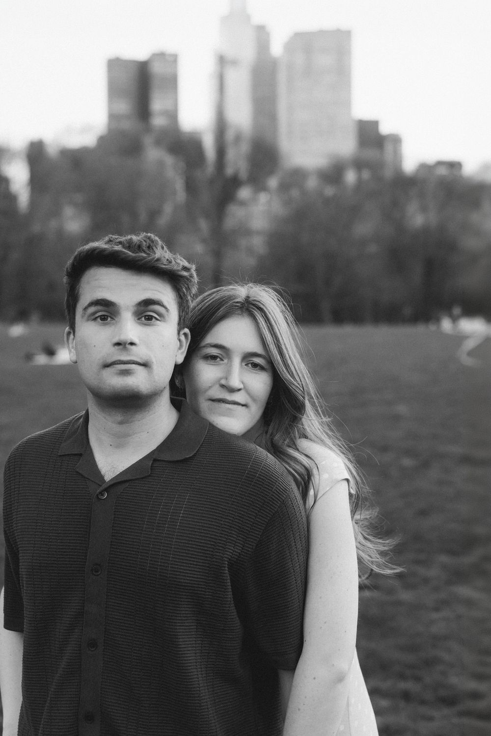 Black and white photo of a young couple posing in a park with city skyline in the background.