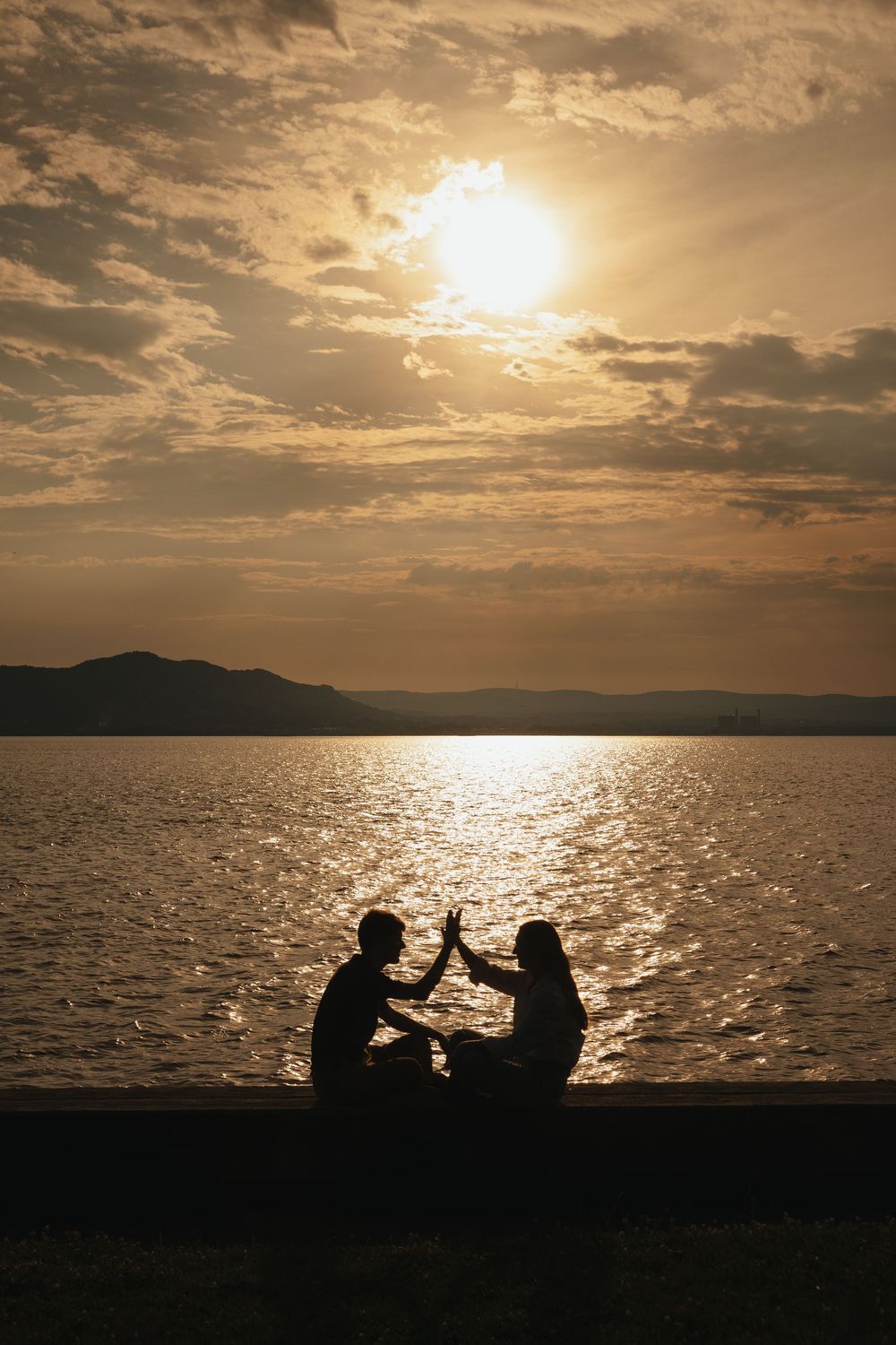 Silhouette of couple toasting drinks by a shimmering lake at golden sunset with mountains in background.