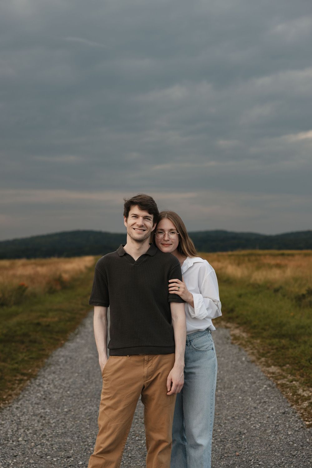 A young couple stands on a gravel path in an open field under a cloudy sky, smiling and embracing.