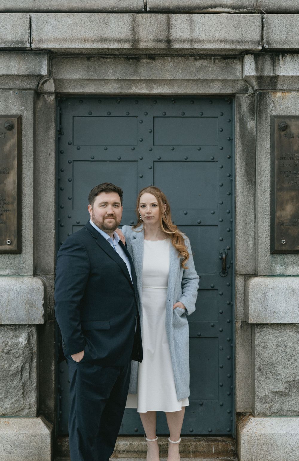 A couple poses in front of a grand stone building with a dark green door, man in navy suit and woman in white dress.