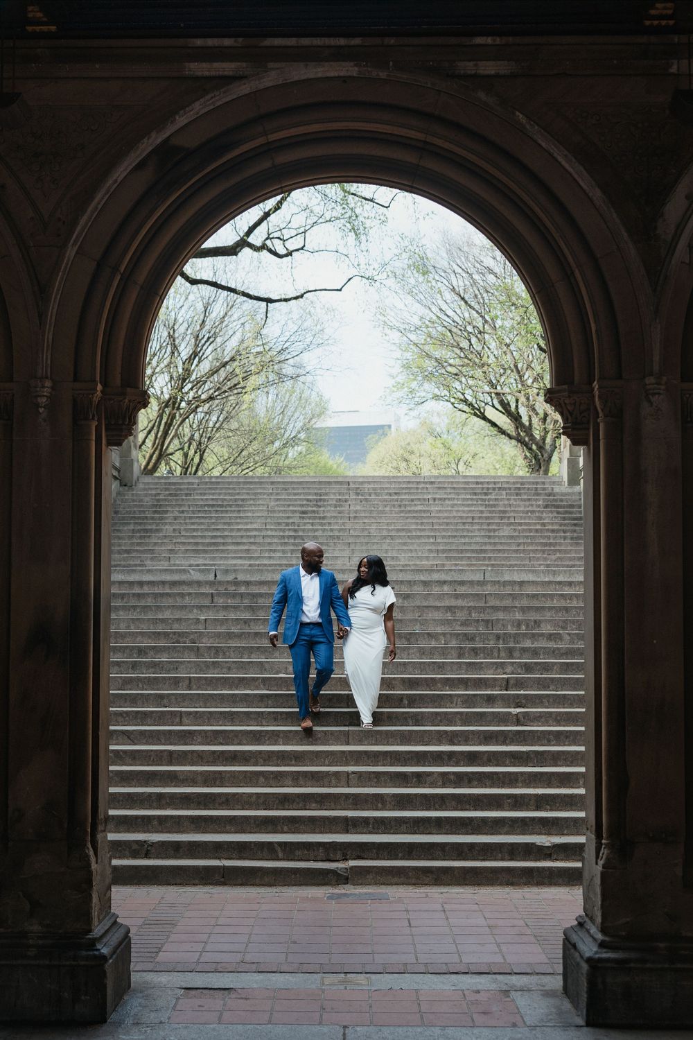 Couple in formal attire walking down stone steps under an arched entrance at Bethesda Terrace in Central Park.