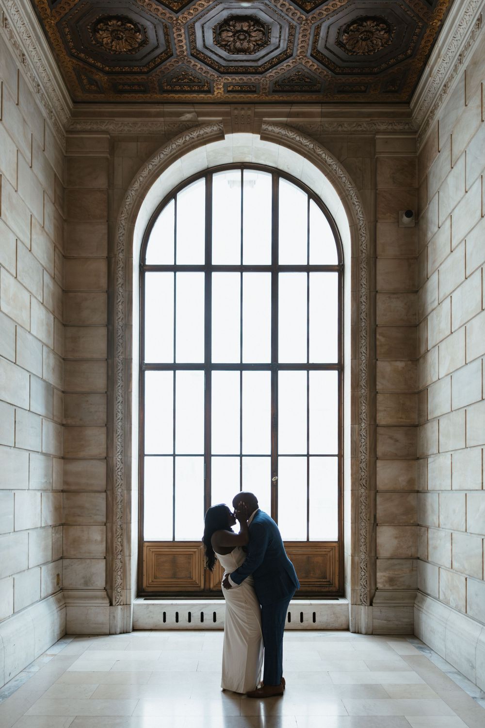 Couple sharing a romantic kiss silhouetted against an arched window in an elegant stone building.