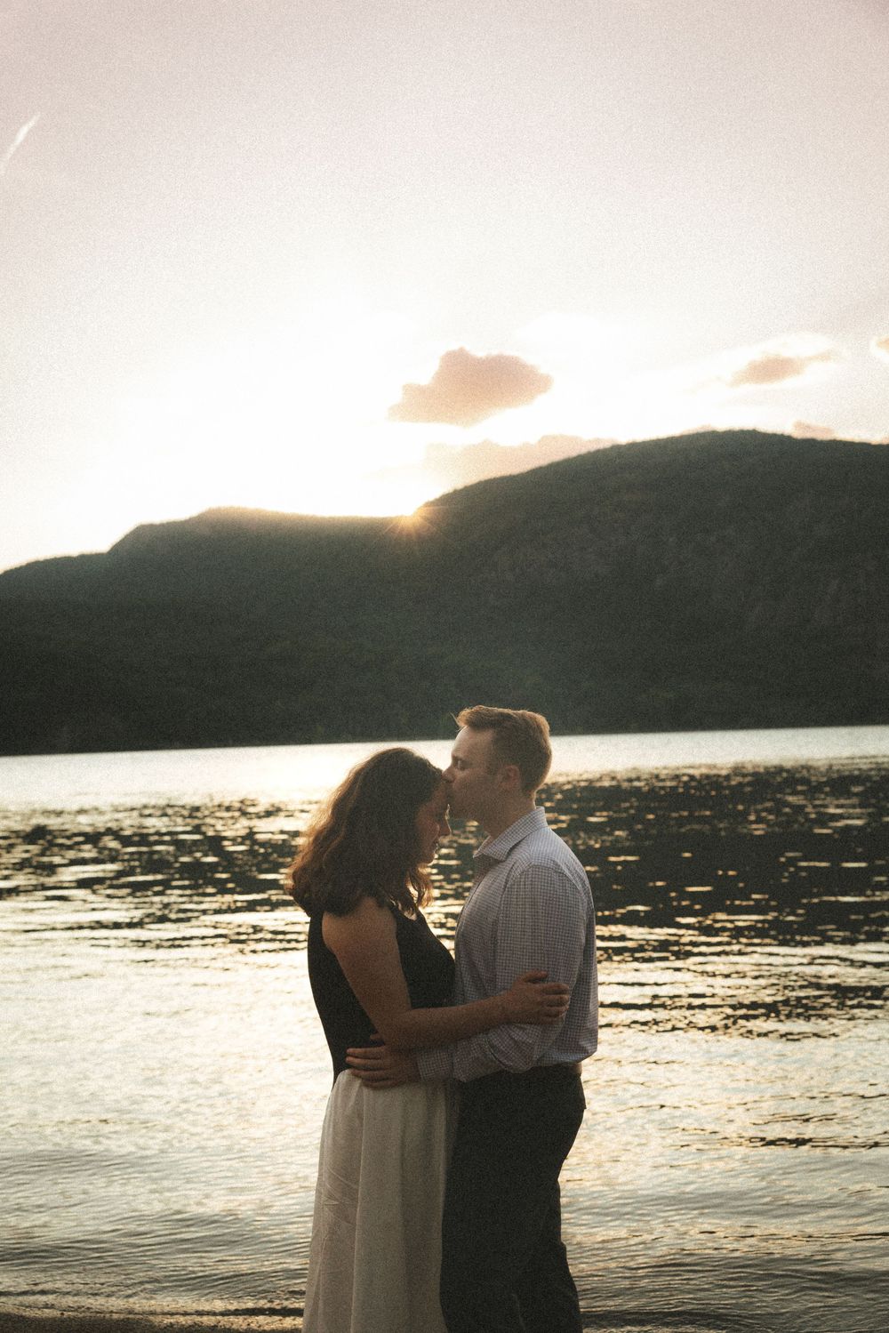 Couple embracing at sunset by a serene lake with mountain silhouette in the background.