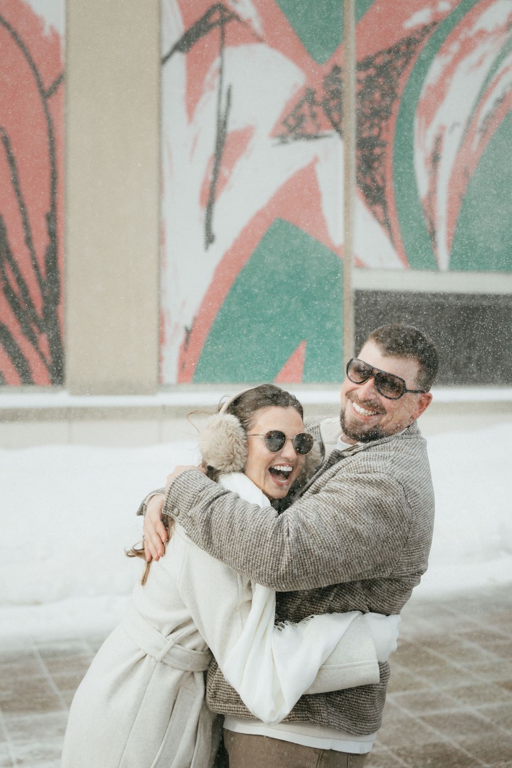 Two people in casual winter attire embrace and laugh together against a white and green geometric wall background.