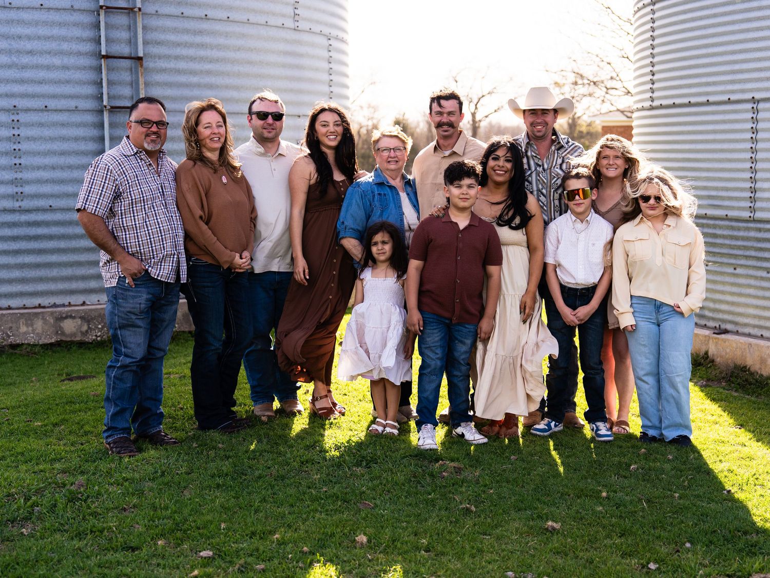 A group of people pose together in front of white farm silos on a grassy area during golden hour.