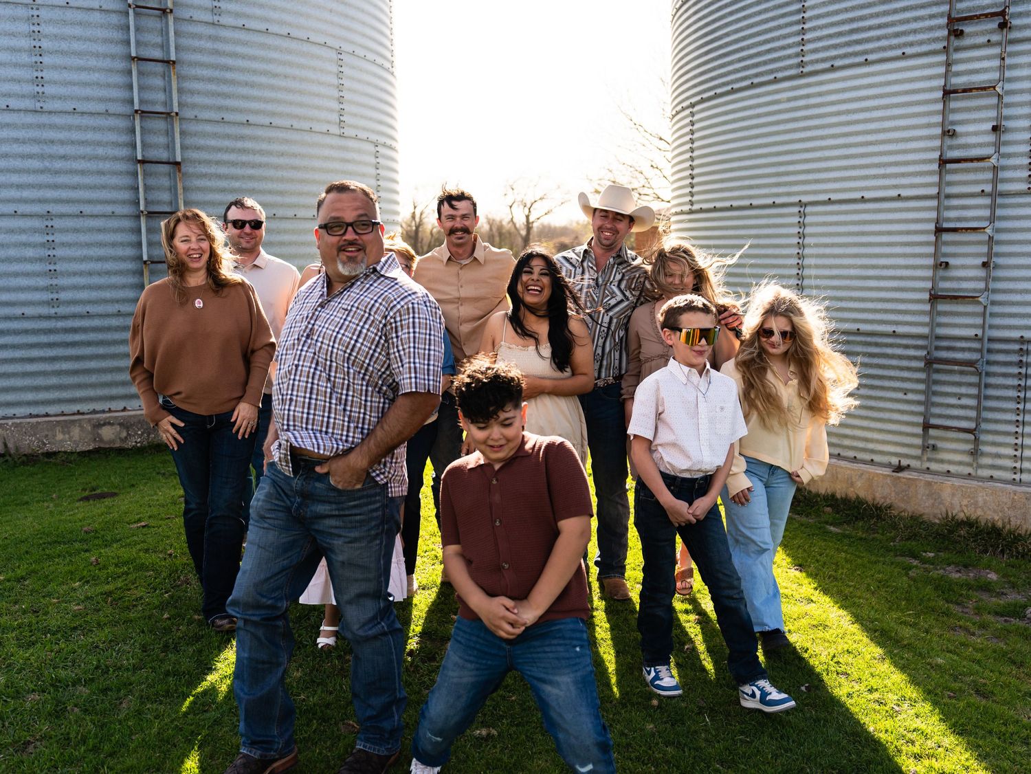 Group posing for photo in front of large grain storage silos on grassy area during golden hour lighting.