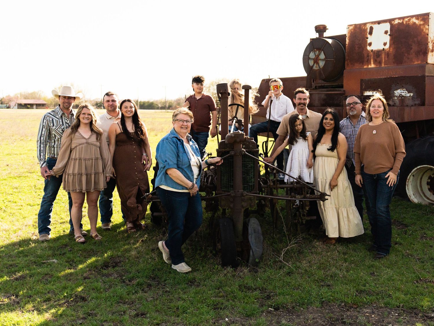 A group of people in rustic clothing pose together near a vintage train locomotive in a grassy field during sunset.