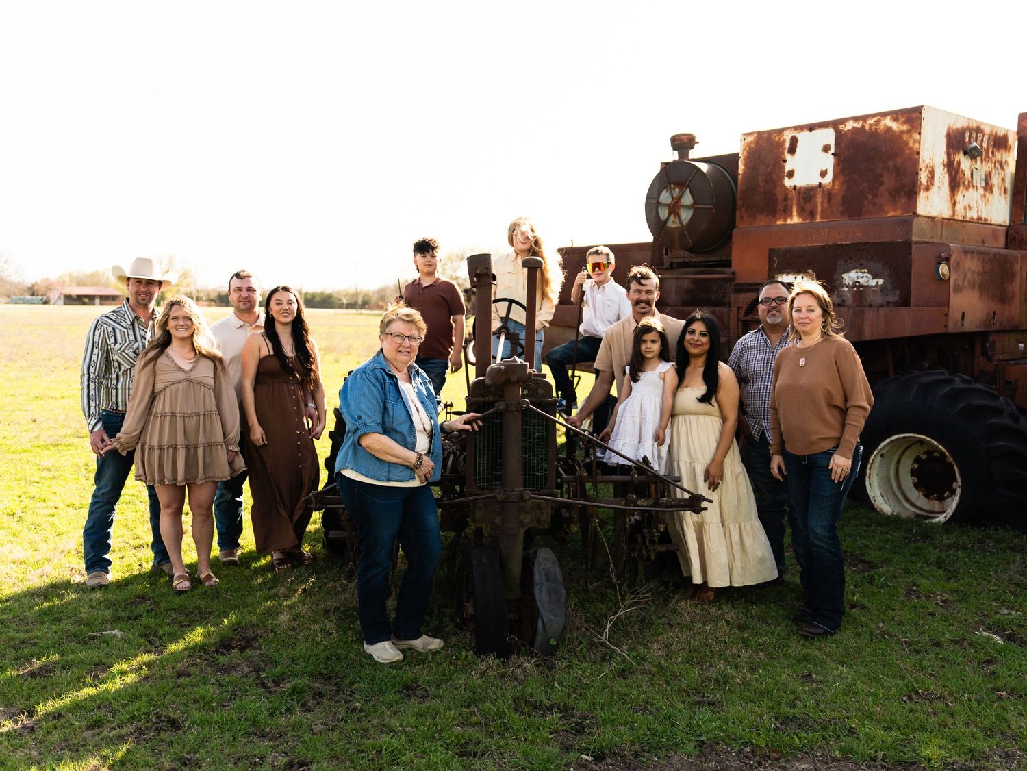 A group of people stand together outdoors in a country setting with an old rustic tractor in the background at sunset.