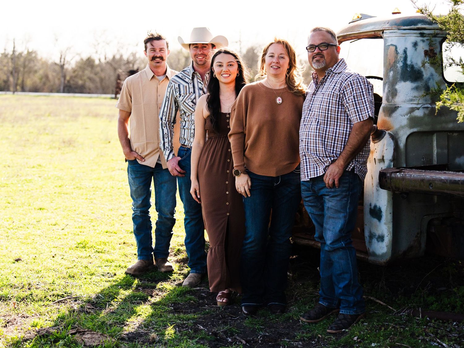 A group of people stand together smiling next to a vintage pickup truck in a sunny field.