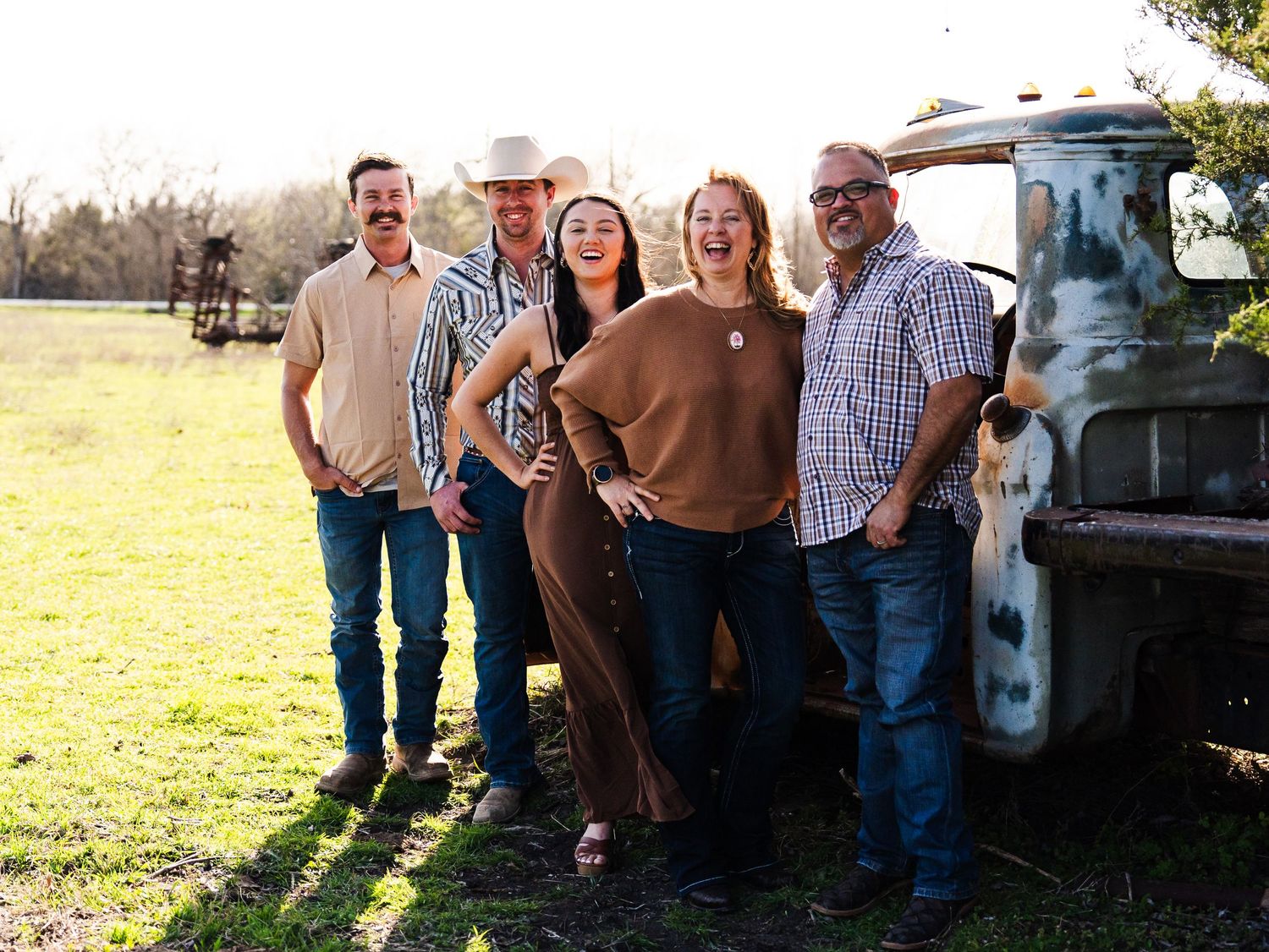 Group of people leaning against vintage pickup truck in sunny field with long autumn shadows.