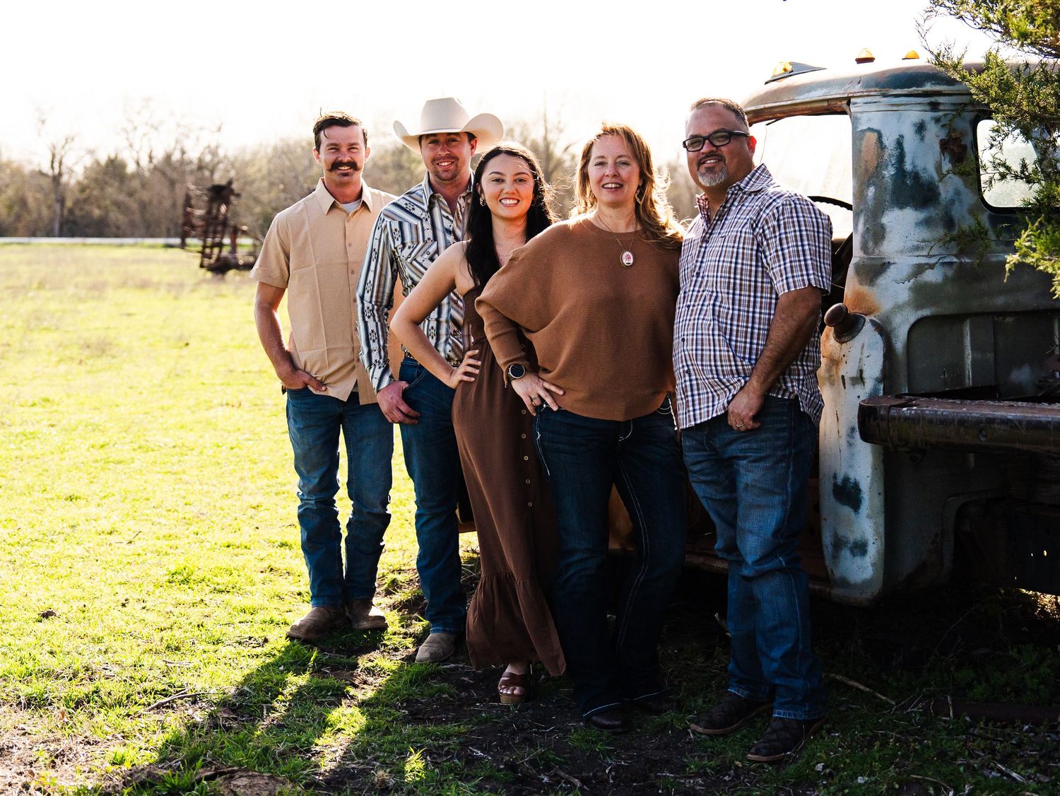 A group of people stand together smiling by an old vintage truck in a sunlit field at golden hour.