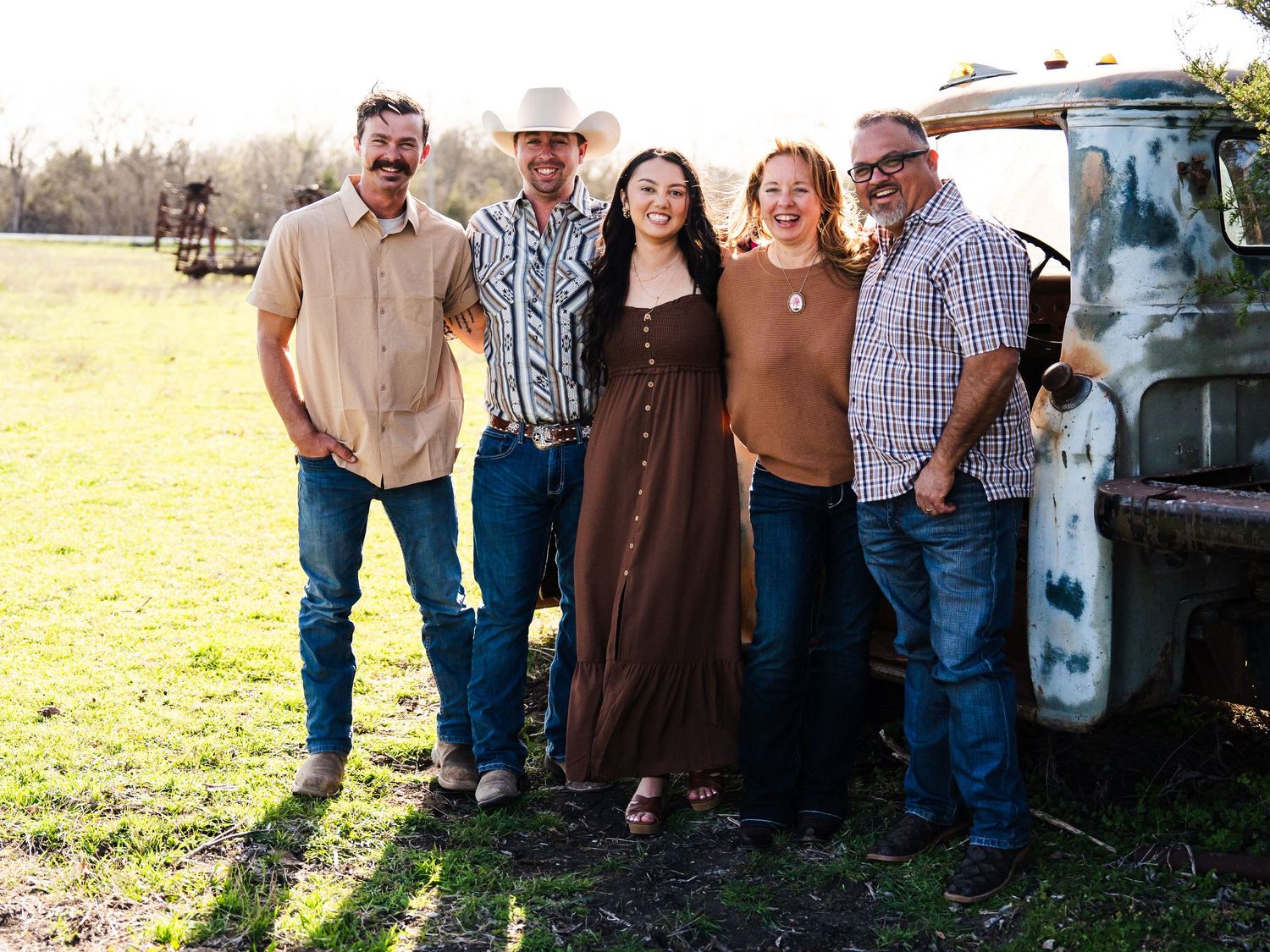 A smiling group in casual clothing standing together outdoors near an old truck with a farm field in the background.