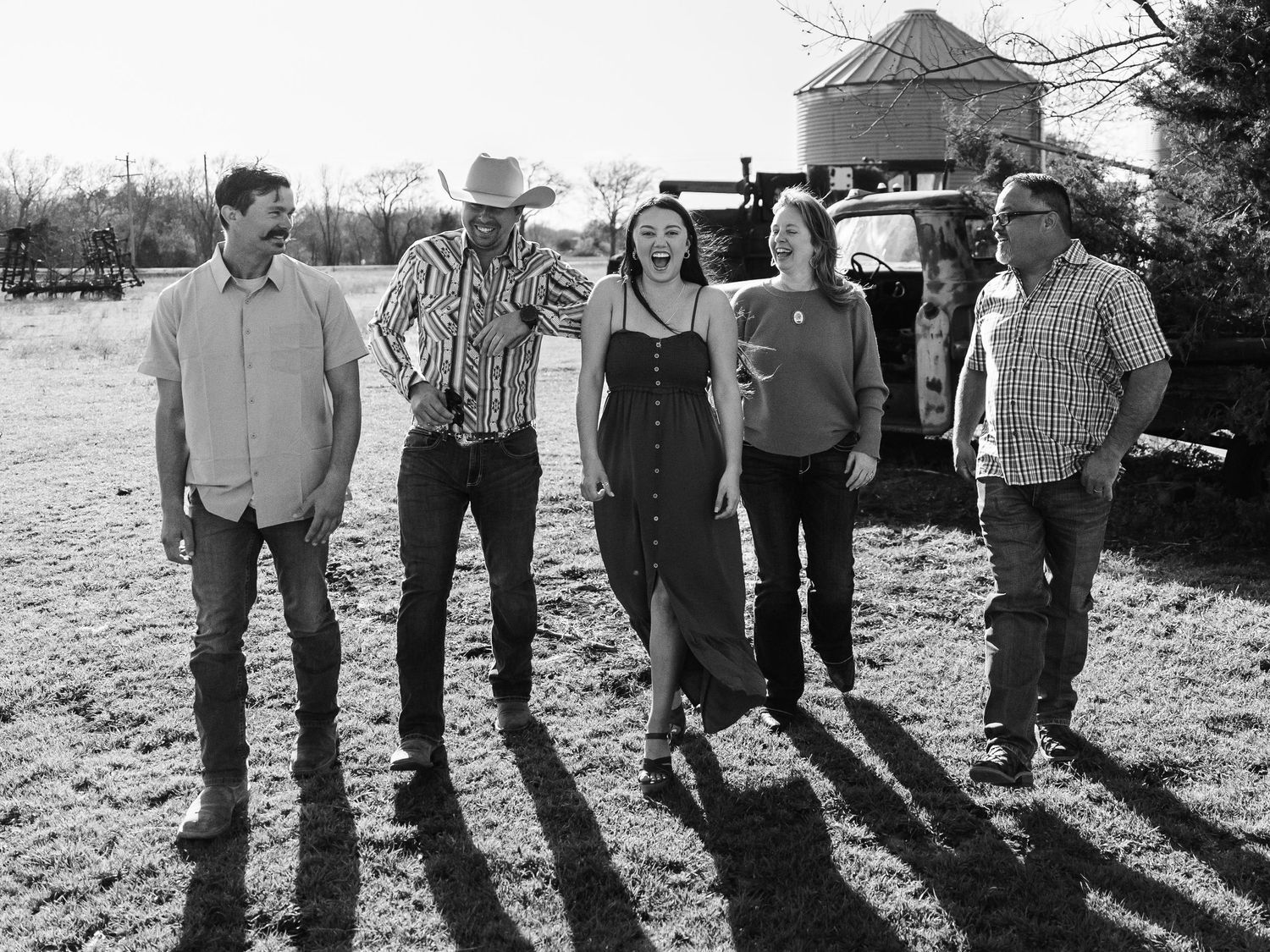 Black and white photo of five people standing in a line casting long shadows on rustic farmland with a building in background.