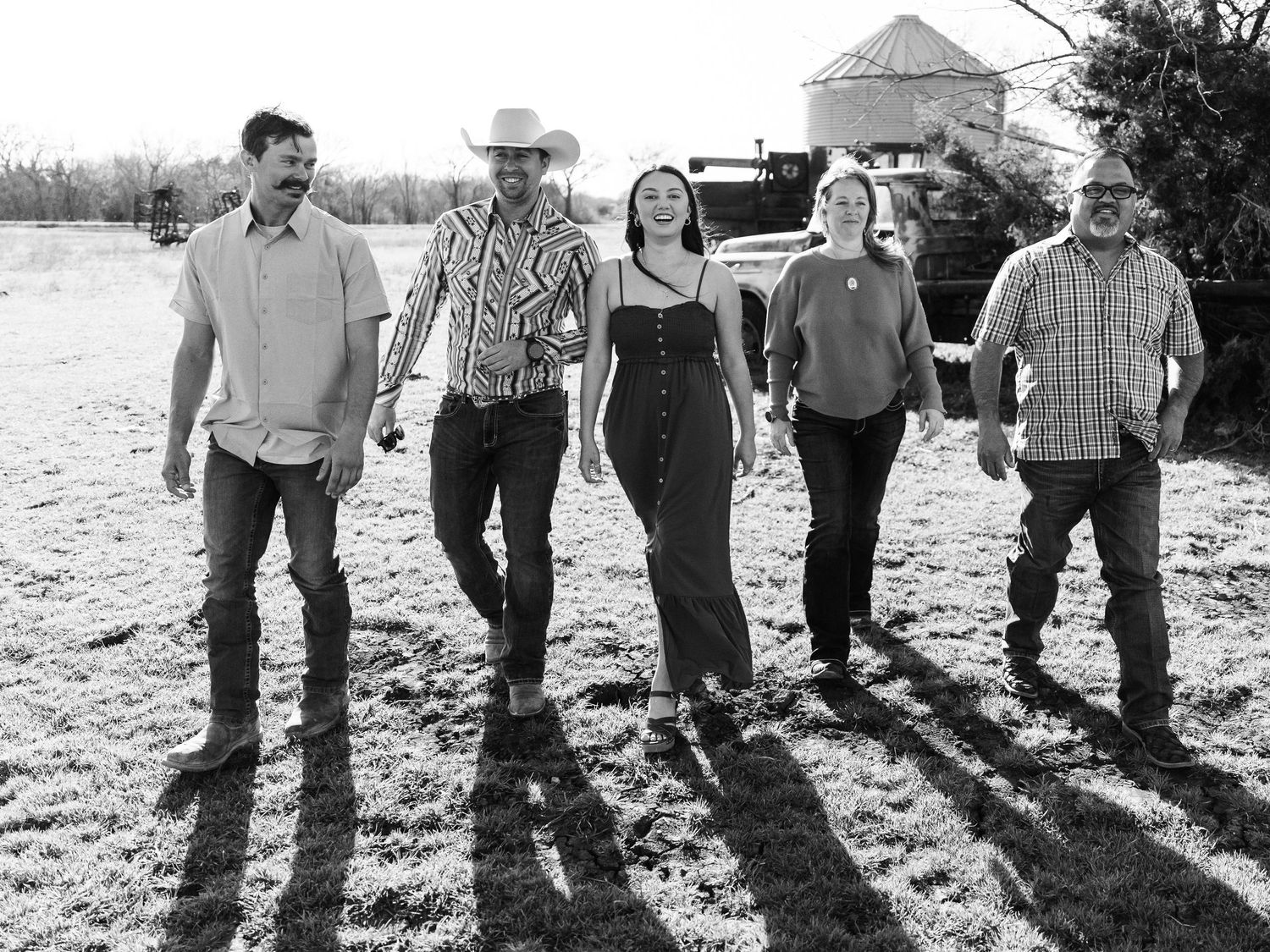 Black and white photo of five people standing in a row casting long shadows on a rural farm landscape.