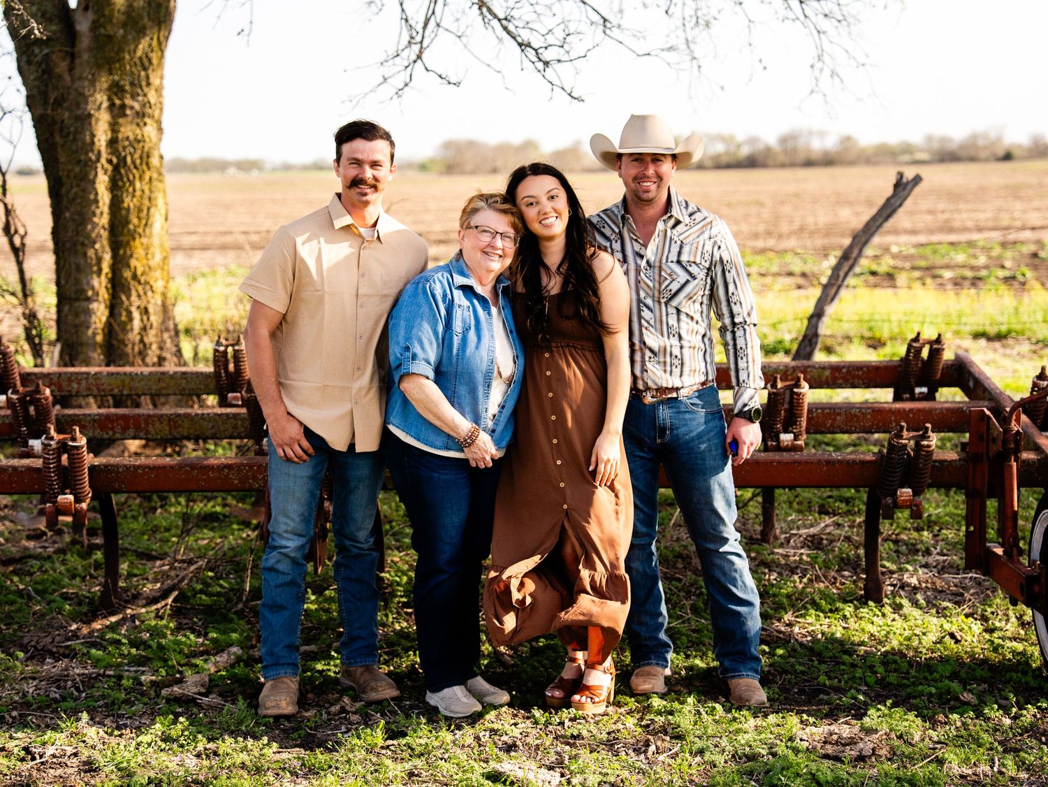 A group stands together outdoors on a farm with rustic farm equipment and a bare tree in the background.