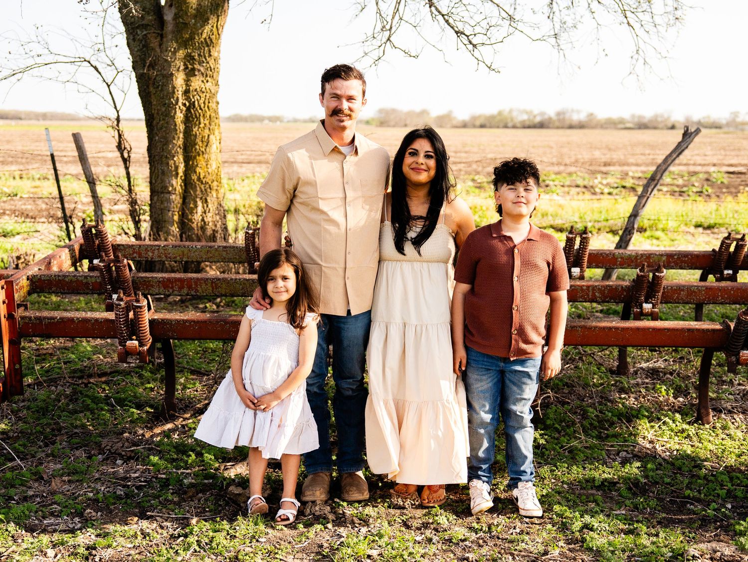 Family of four poses together outdoors in front of wooden benches and a scenic country field on a sunny day.