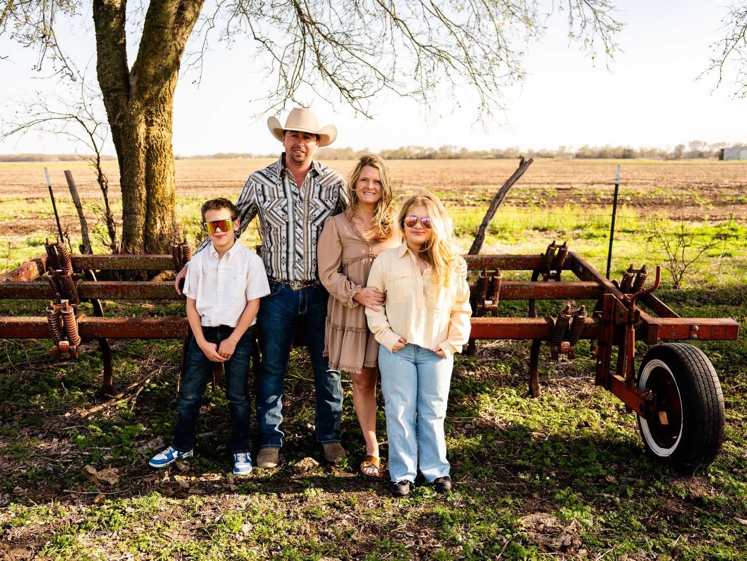 A group stands together on a rural farm setting with an old wheelbarrow and wooden fence in the background.