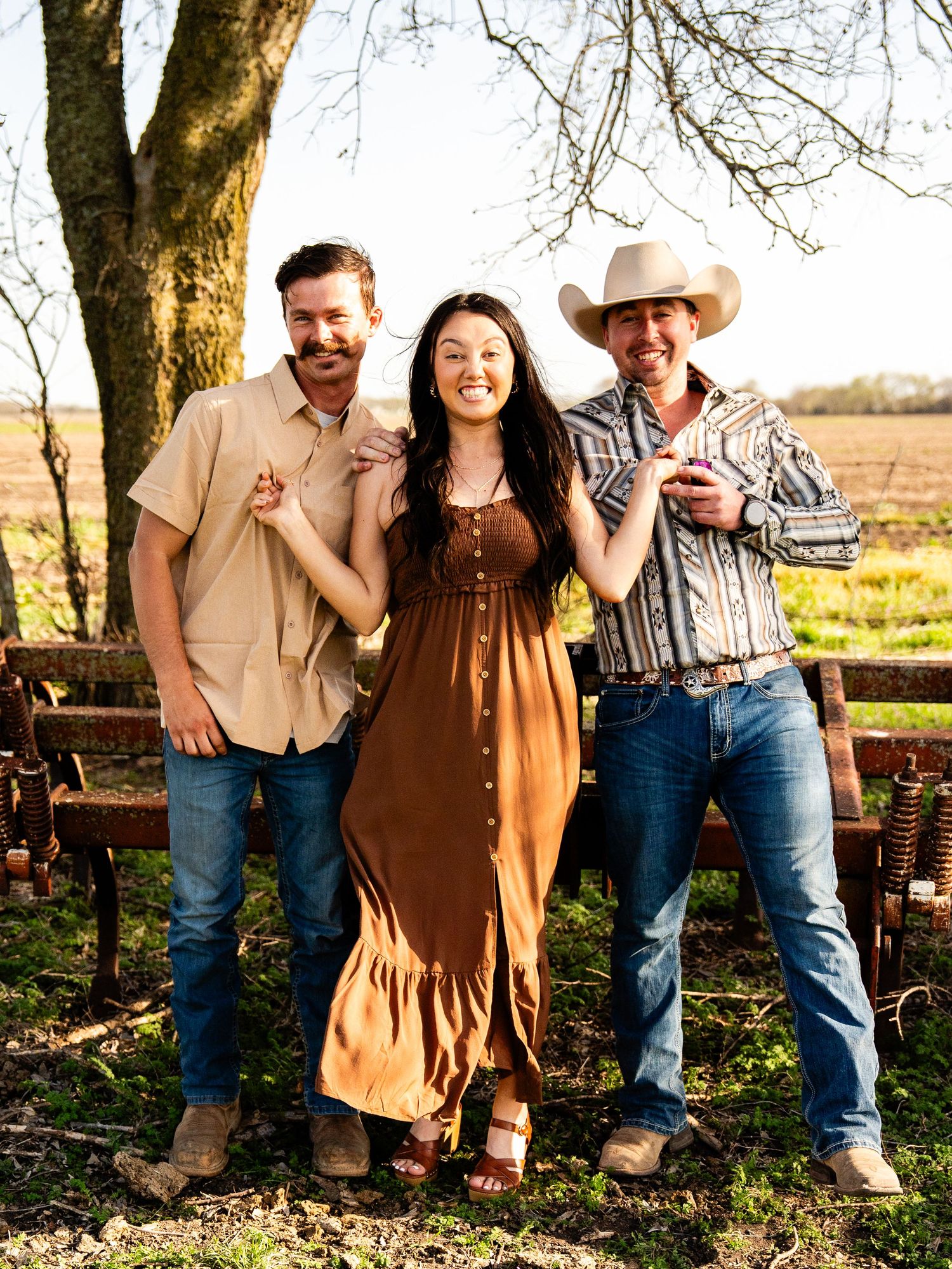 Three people in western-inspired outfits pose together outdoors near a wooden fence on a sunny day.