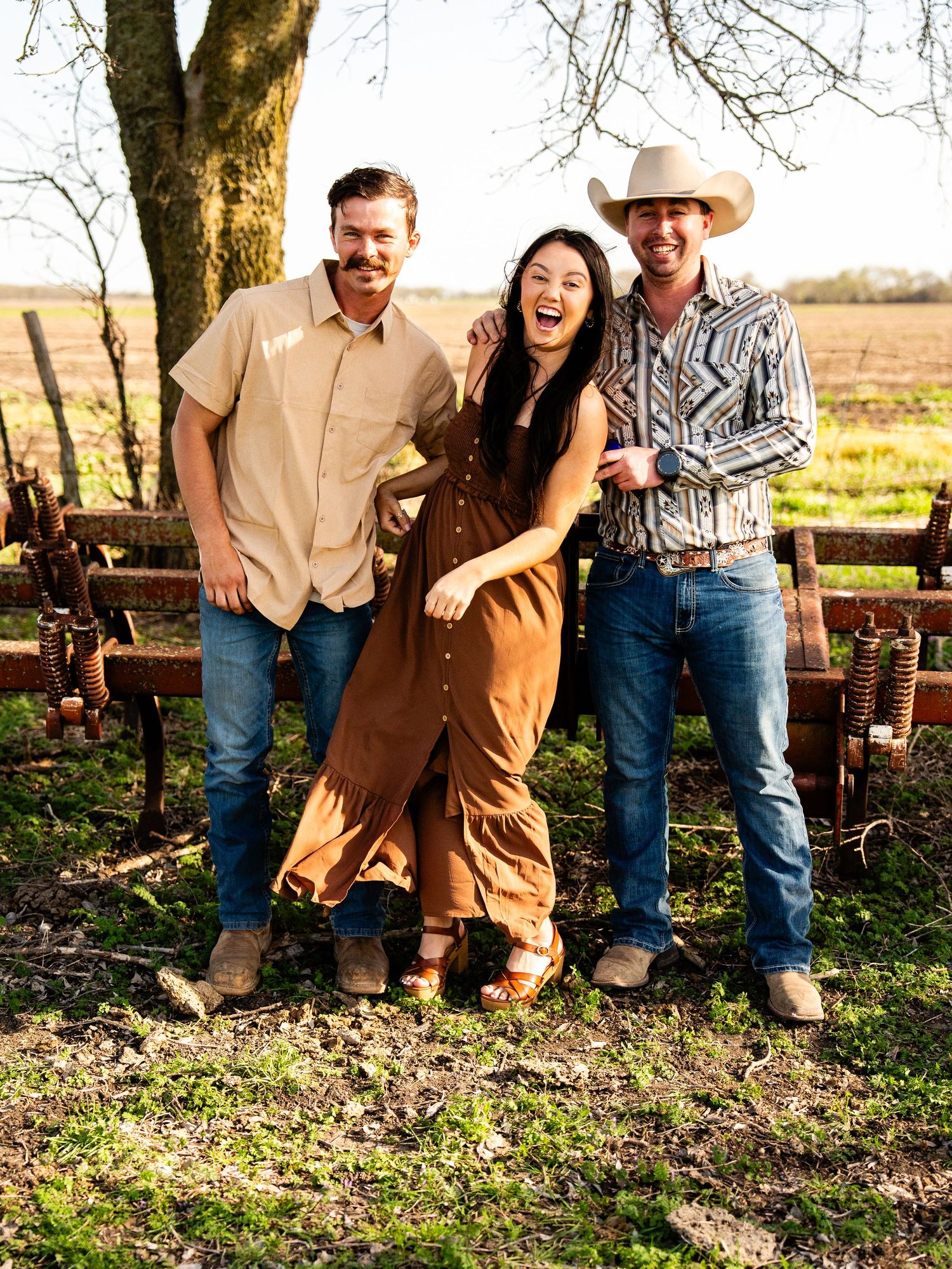 Three friends share laughs together while posing for a casual photo on a rustic ranch with a wooden fence background.