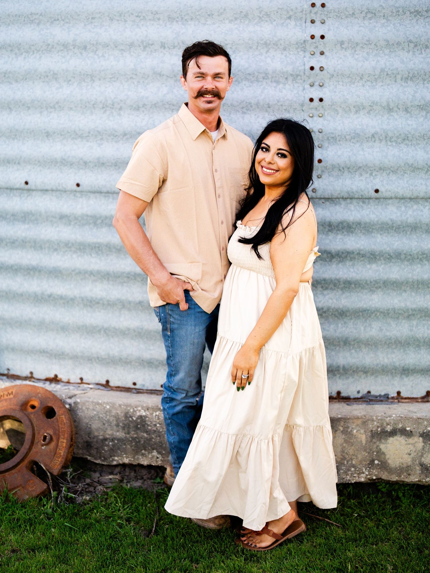 A couple poses together against a corrugated metal wall wearing casual attire with a rustic outdoor backdrop.