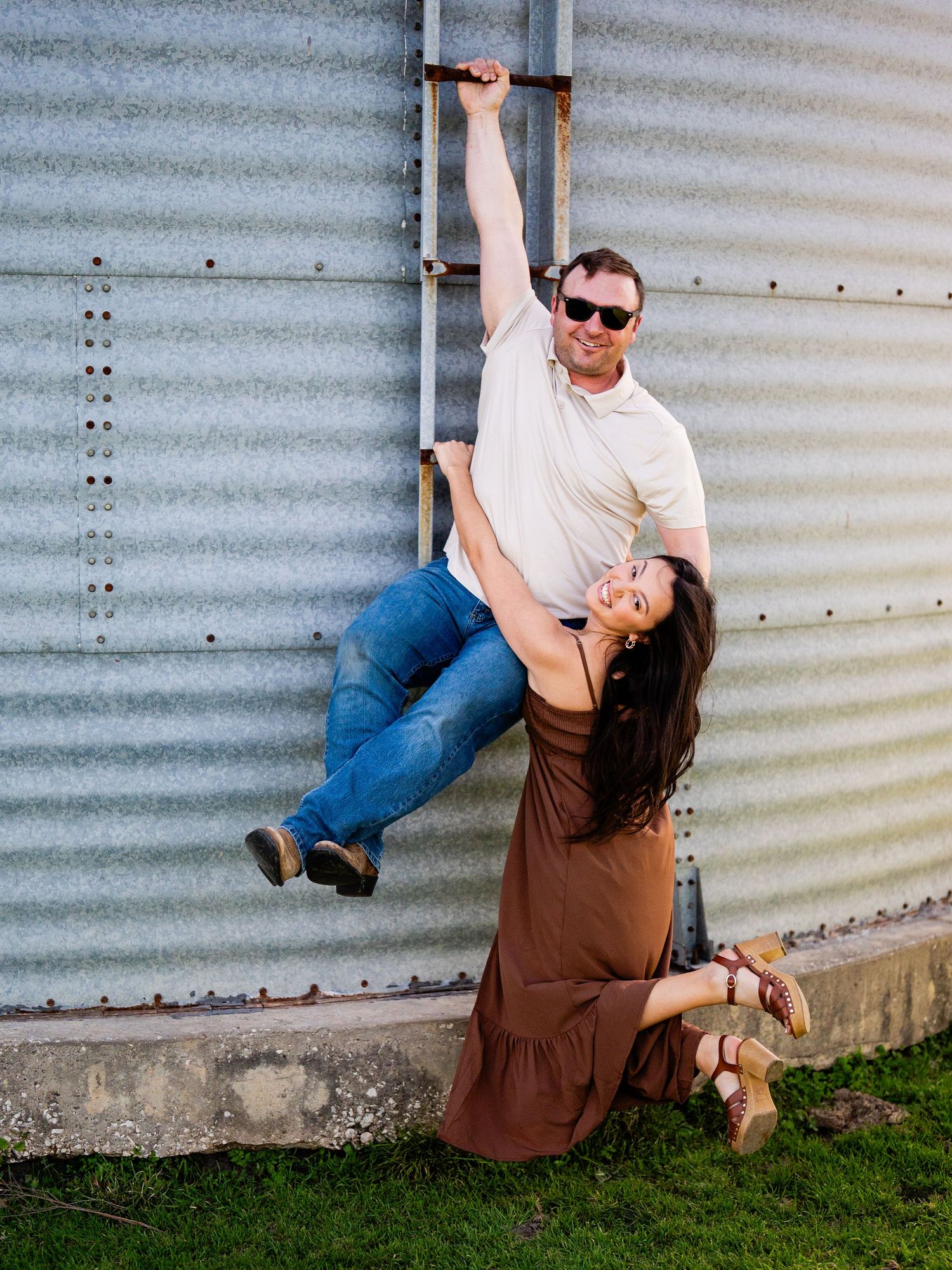 Playful couple poses against corrugated metal wall with one in brown dress and other in jeans and white shirt.
