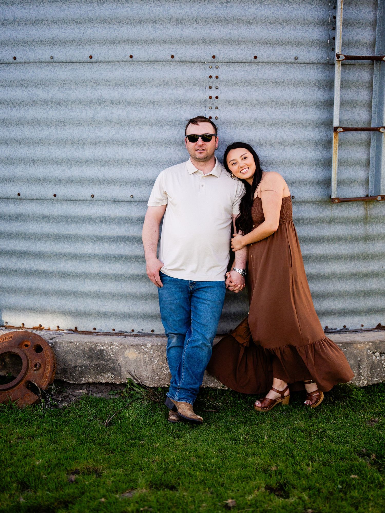 A couple poses together against a corrugated metal wall on a grassy patch on a sunny day.