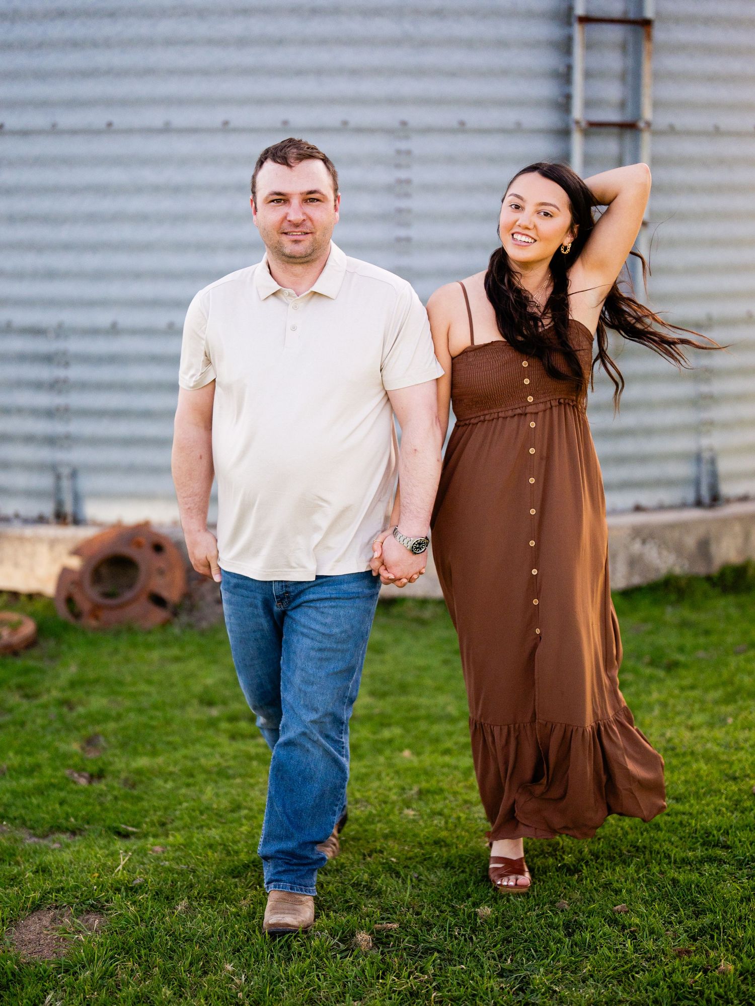 A couple stands together holding hands on grass in front of a metal grain silo on a sunny day.