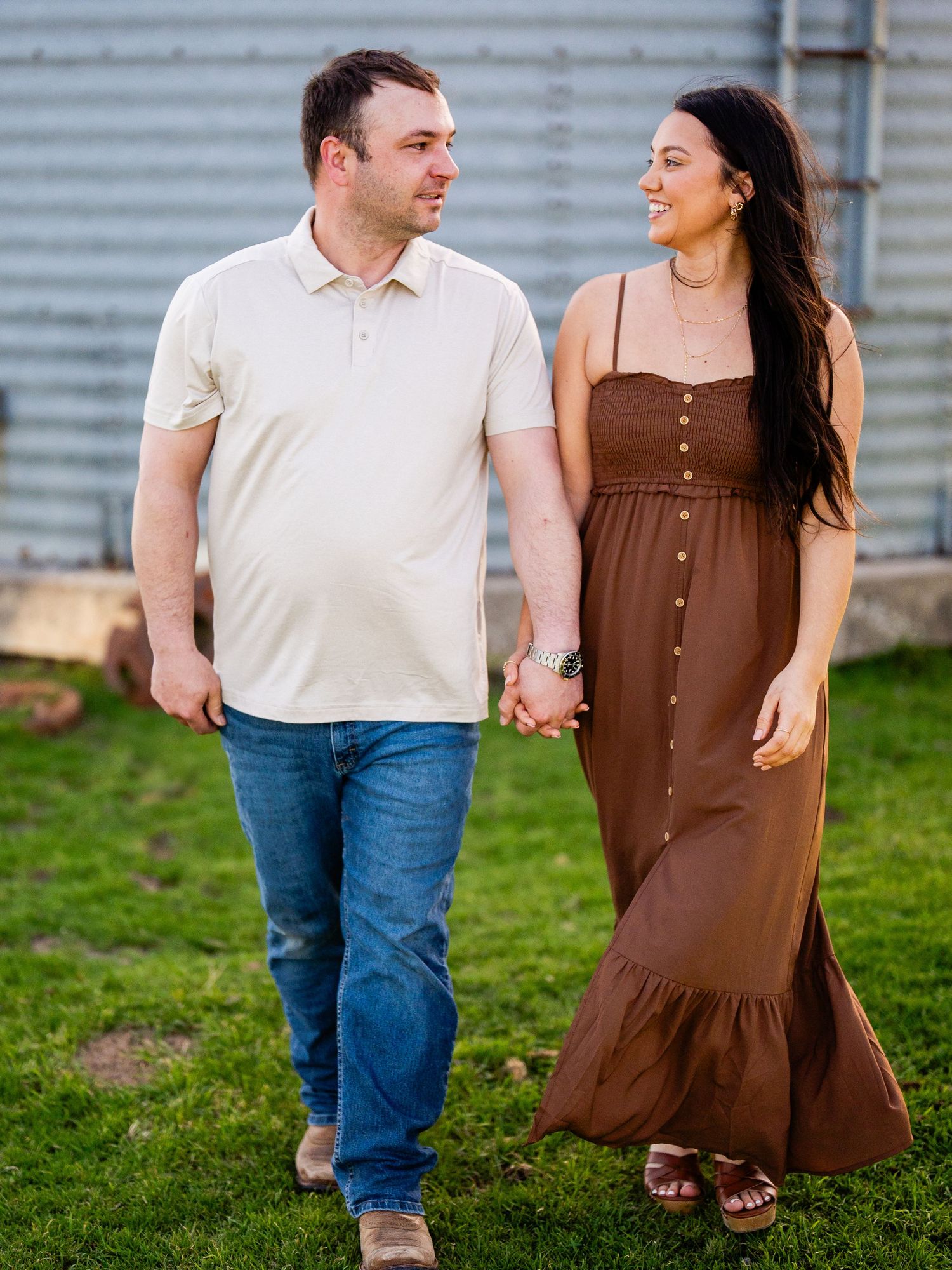 A couple holding hands while walking on grass near a metal wall, with one wearing a brown maxi dress and sandals.