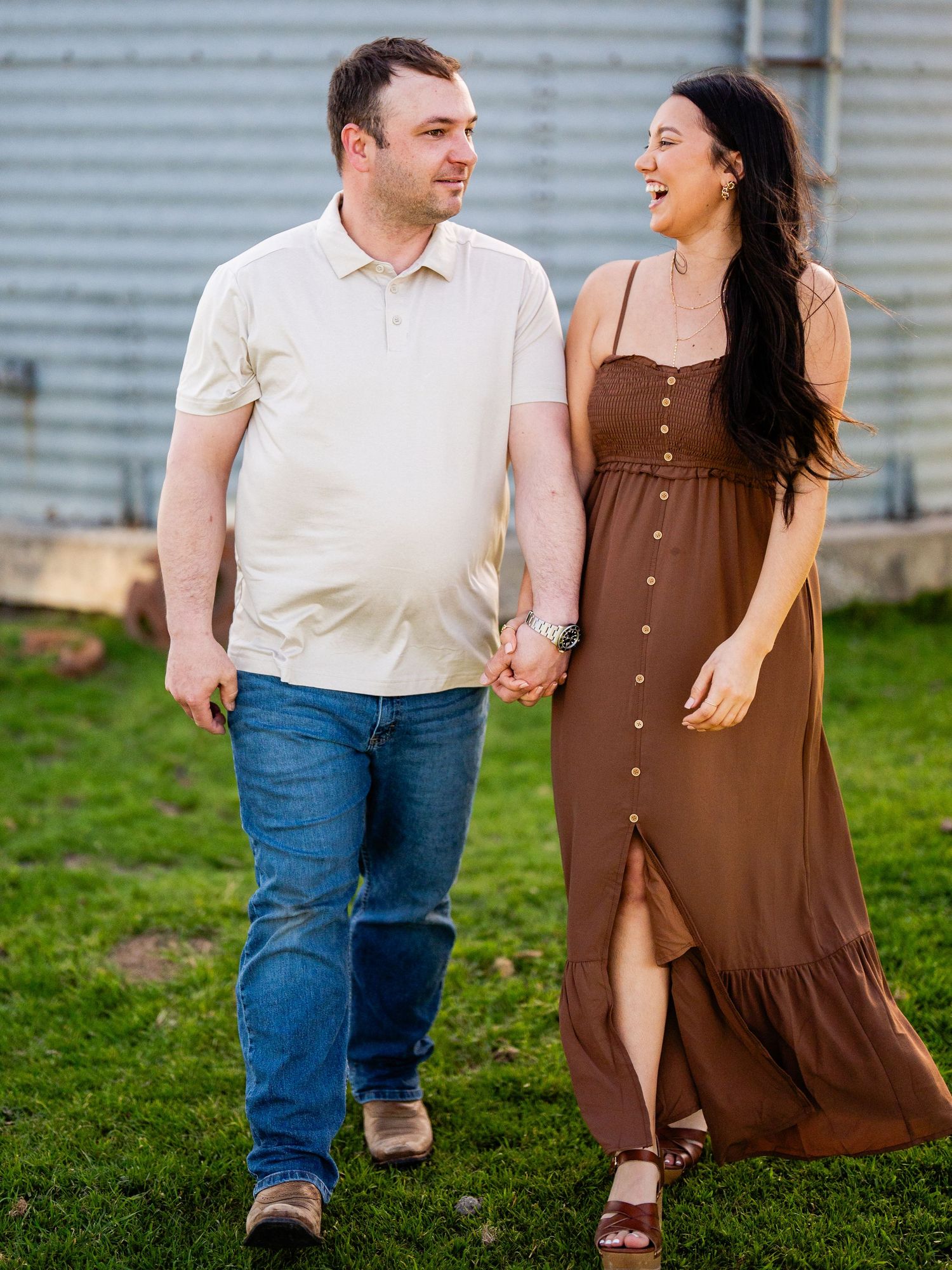 A couple holds hands while walking on grass in front of a corrugated wall, with her wearing a brown dress and him in white.