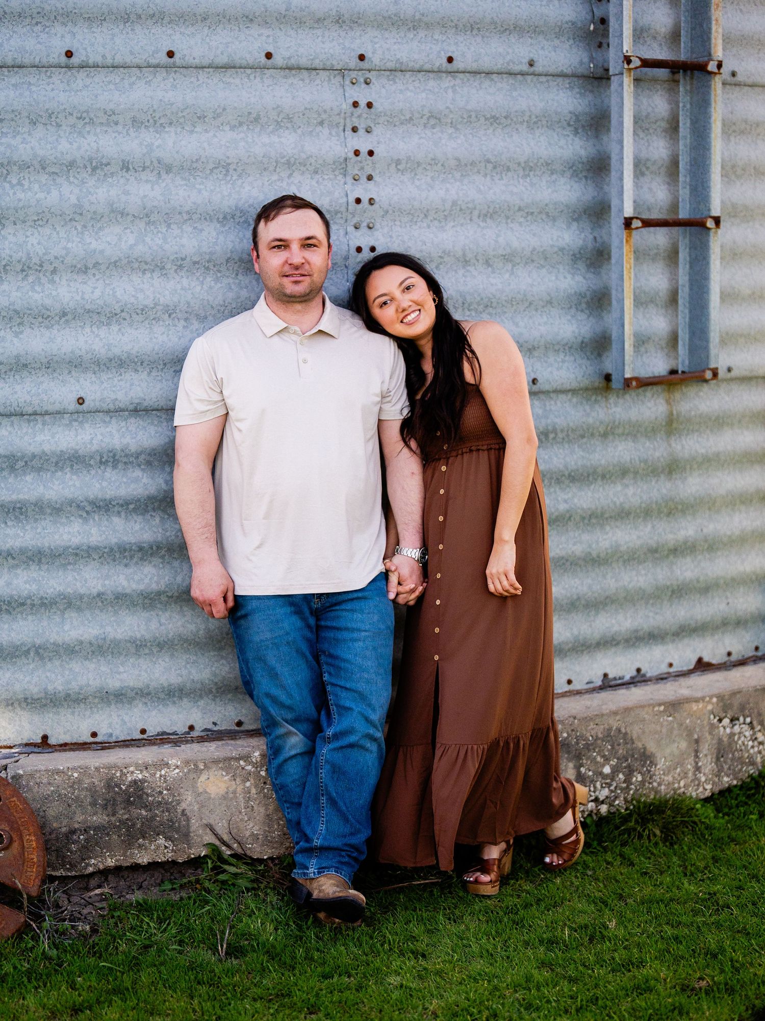 A couple poses together against a rustic metal wall on grass during a casual outdoor photo session.
