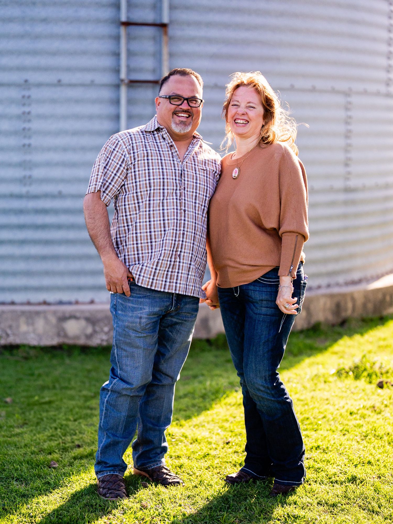 A couple stands together smiling in front of a metal grain silo on a sunny day in casual attire.
