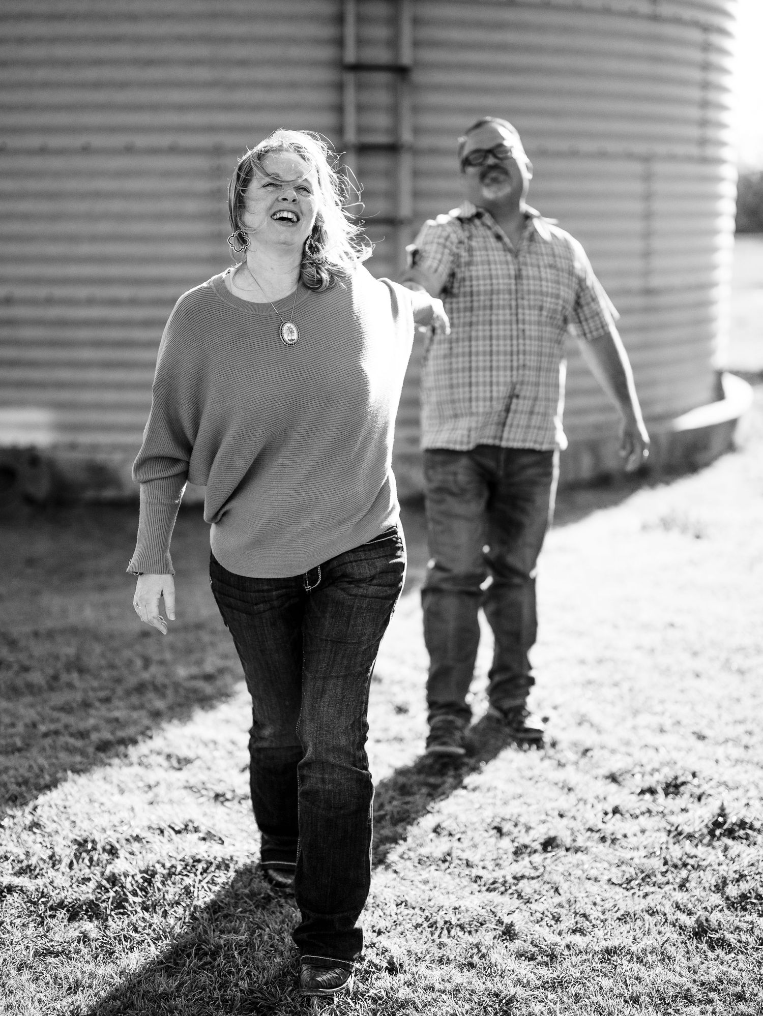 A joyful candid black and white photo of two people laughing together in a rural setting with grain bins in background.