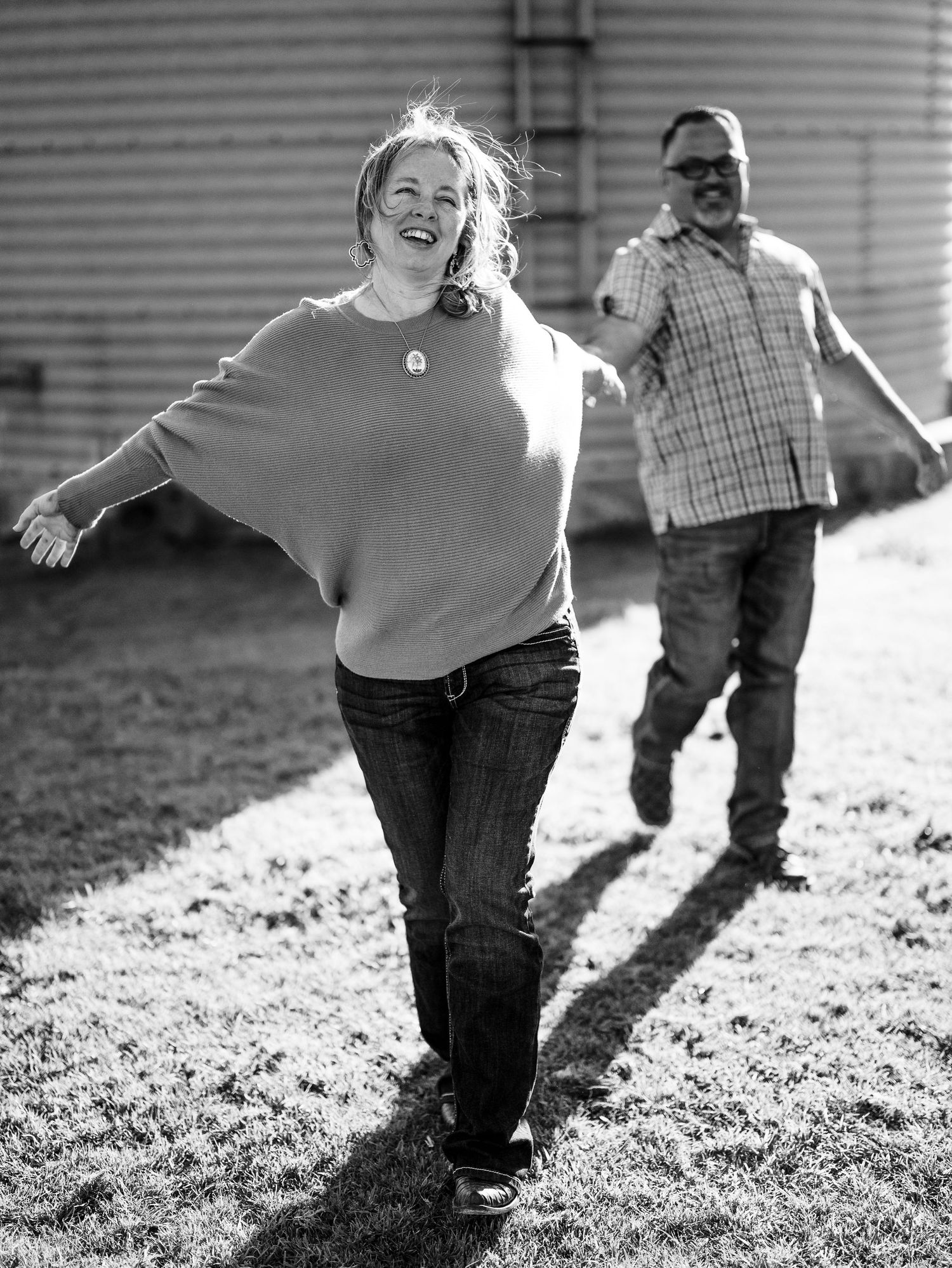 A joyful black and white image shows two people running playfully on grass in front of metal grain silos.