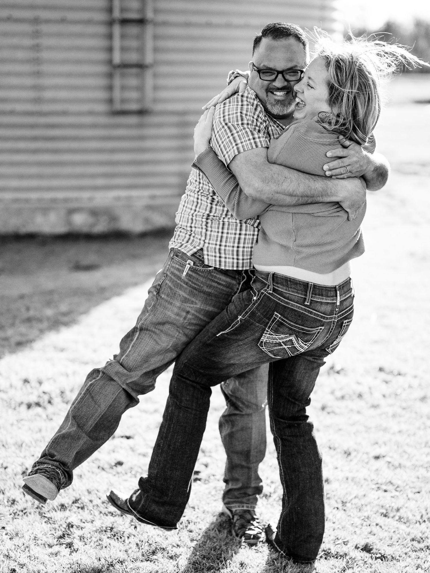 A joyful black and white photo captures a playful embrace between a couple in casual denim and plaid outdoors.