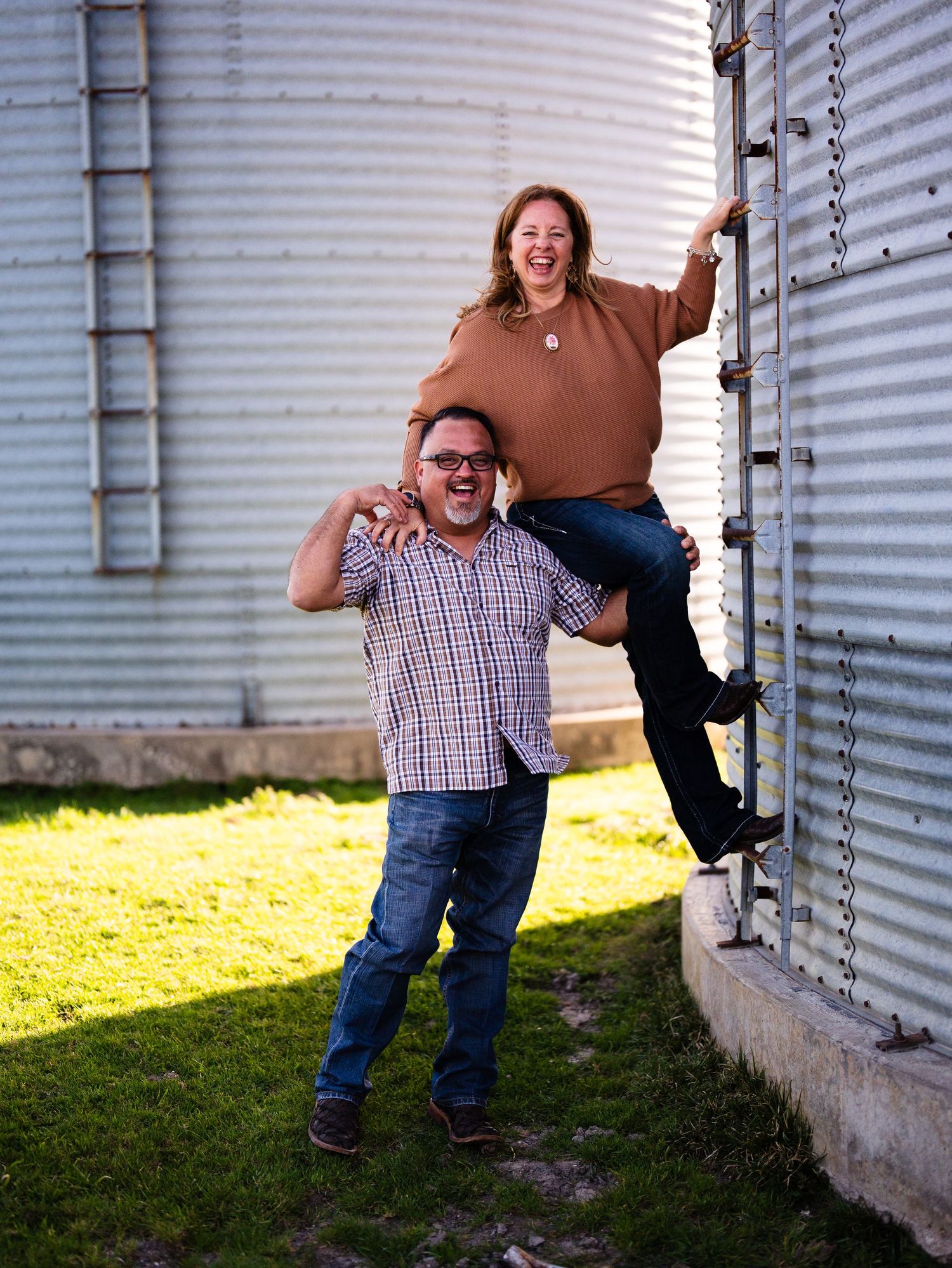 A playful couple poses together near metal grain silos on a sunny day in casual attire.