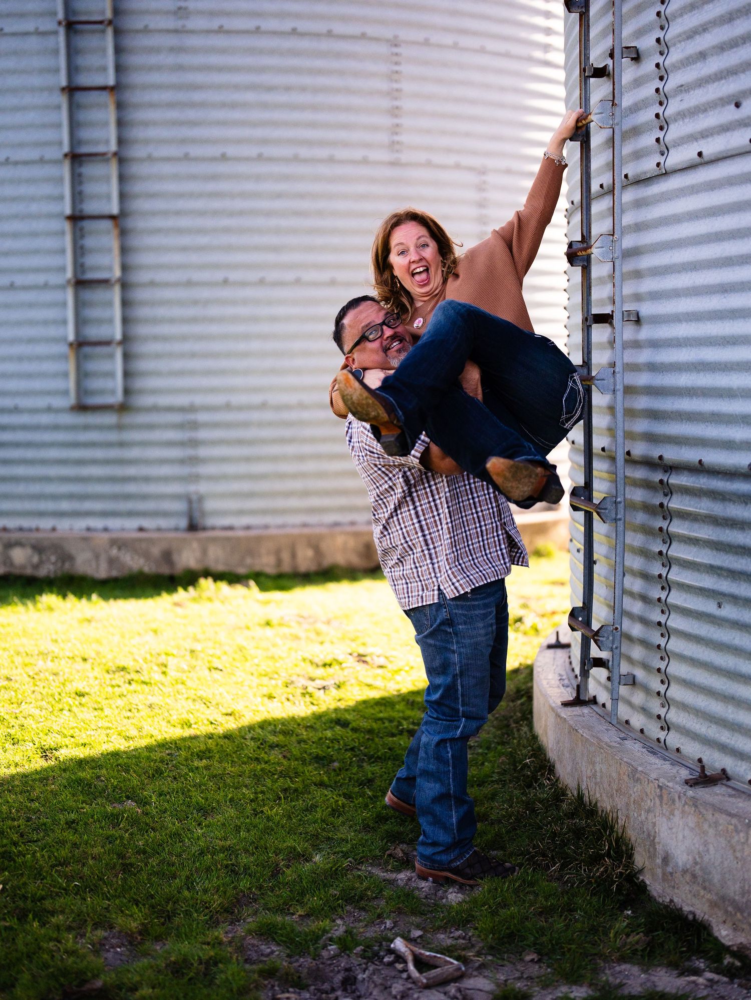 Two people pose playfully by a metal grain silo on a sunny day with green grass in the foreground.