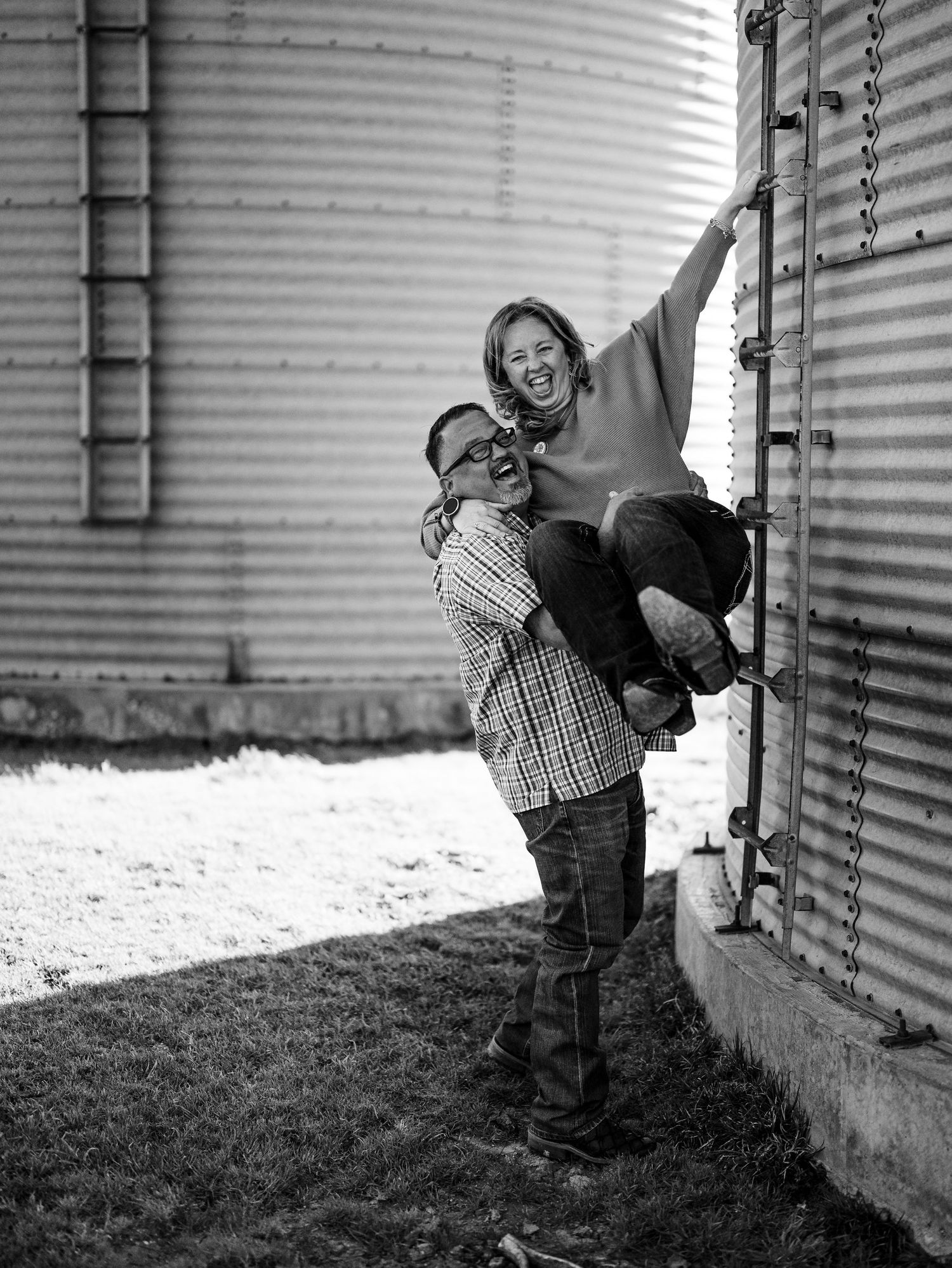 A joyful black and white photograph shows a playful moment between two people near a grain silo in a rural setting.