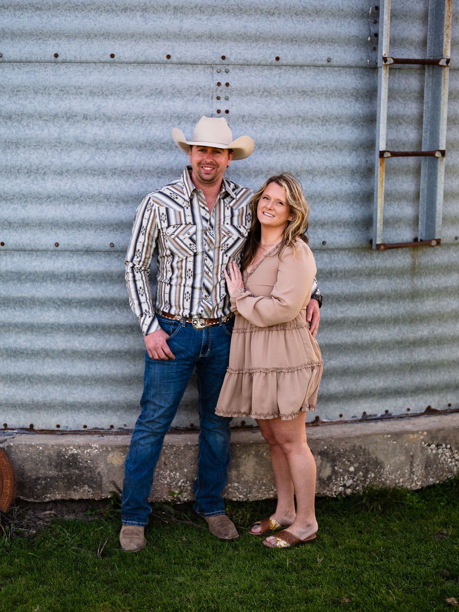 A couple stands together against a corrugated metal wall, with one wearing western attire and the other in a beige dress.