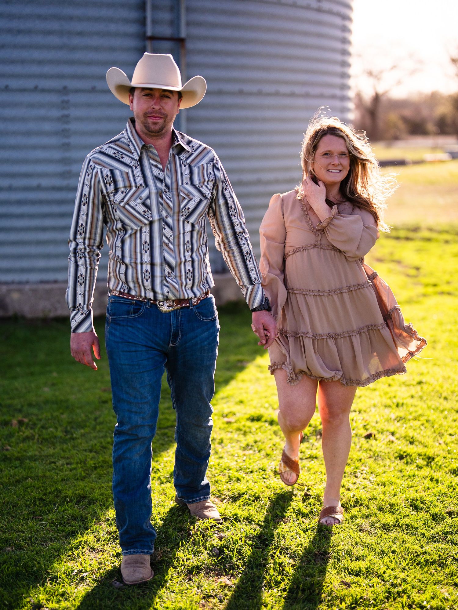 A couple in western wear walks through a sunny field next to a metal grain silo, casting long shadows on green grass.