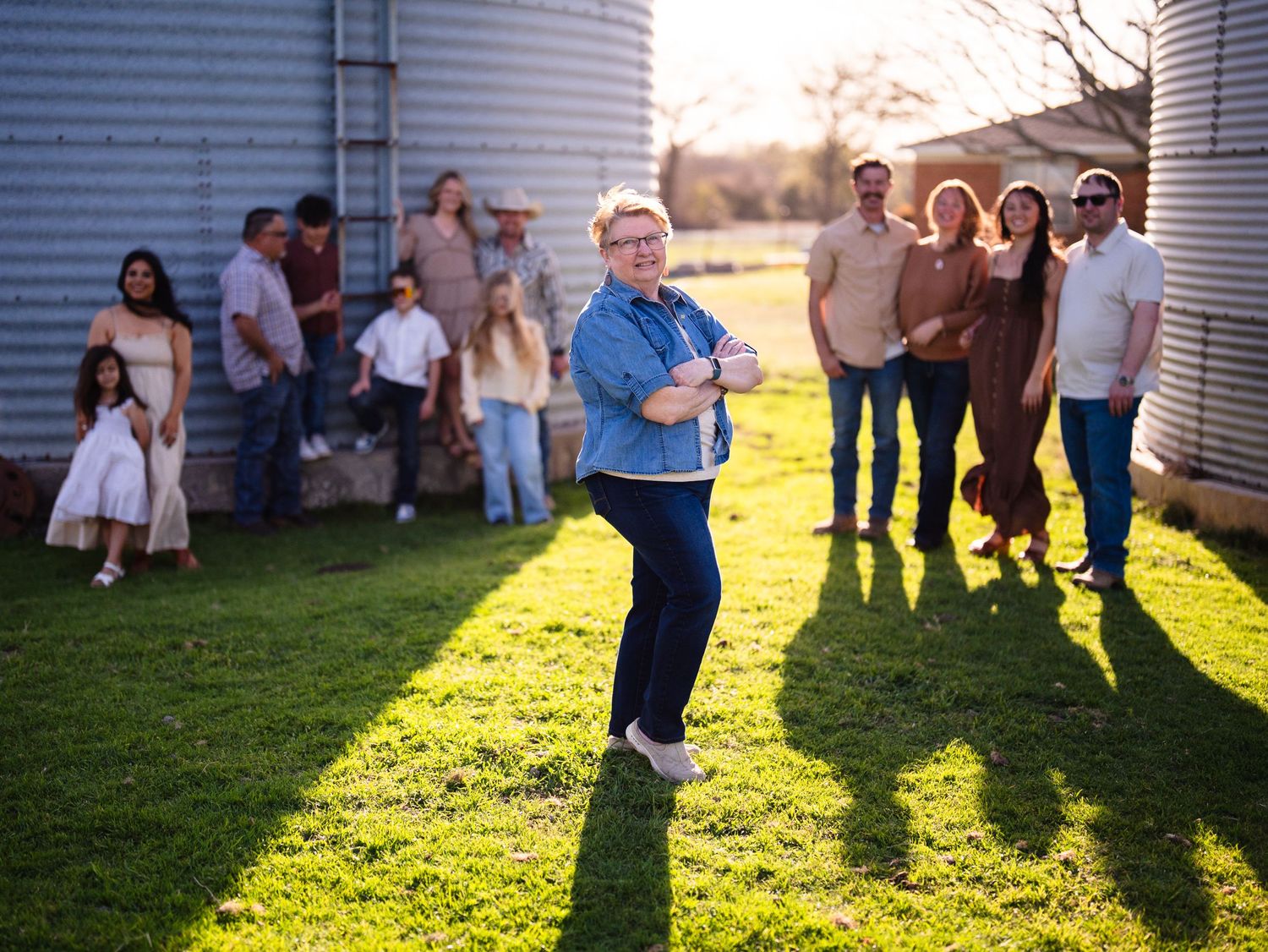 A group of people gathered on grass between grain silos during sunset casting long shadows on a warm evening.