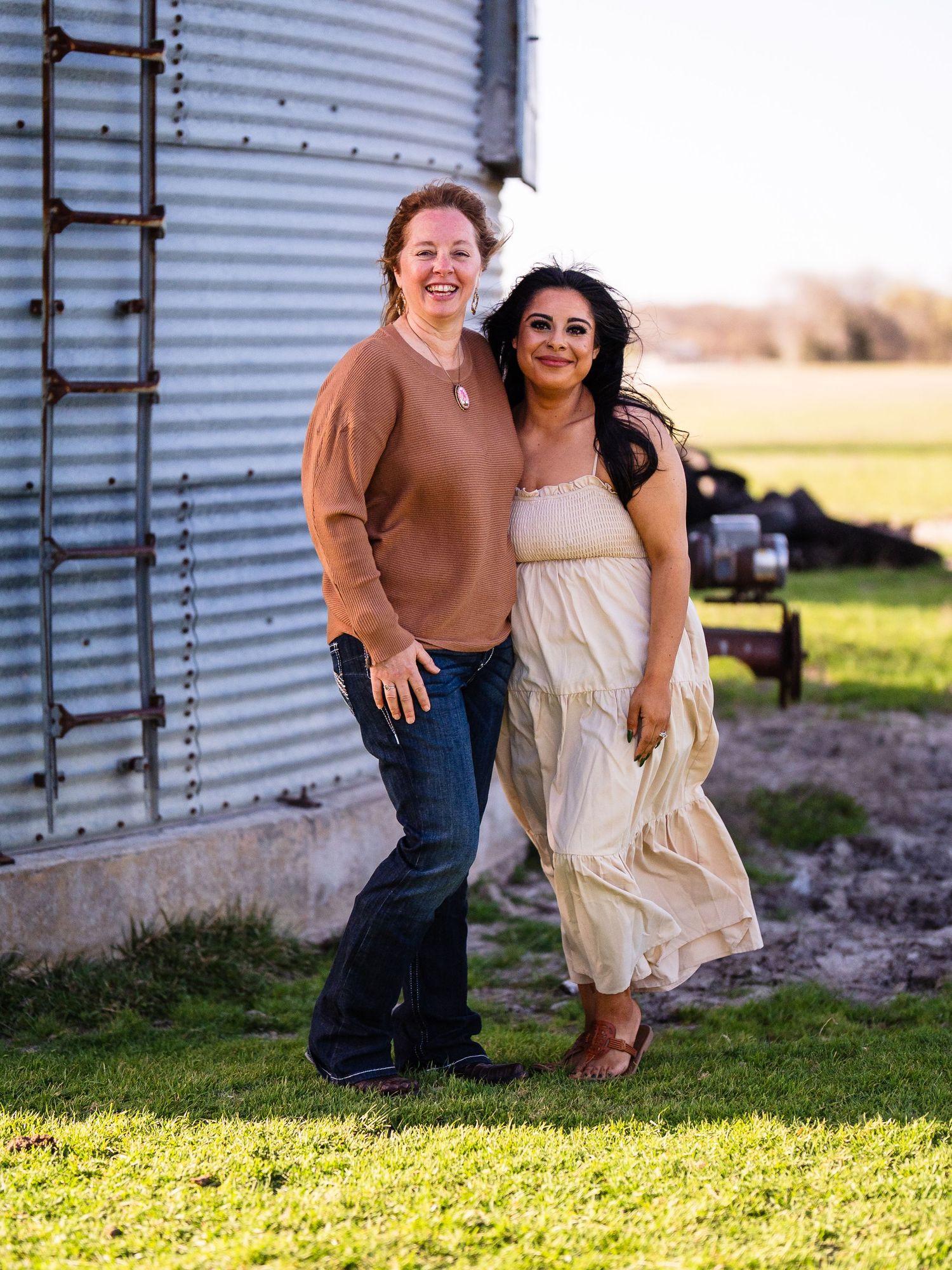 Two friends stand together smiling outside a metal grain silo on a sunny day in the countryside.