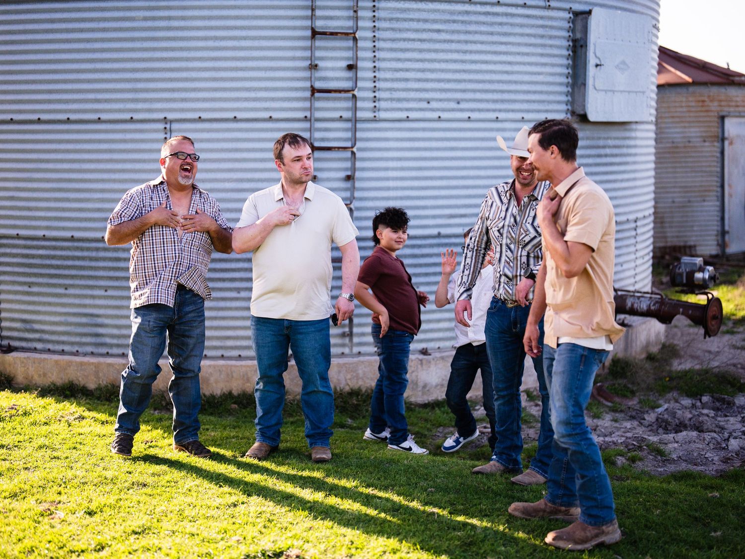 A group of people gathered casually near a metal grain silo on a sunny day, engaged in conversation.