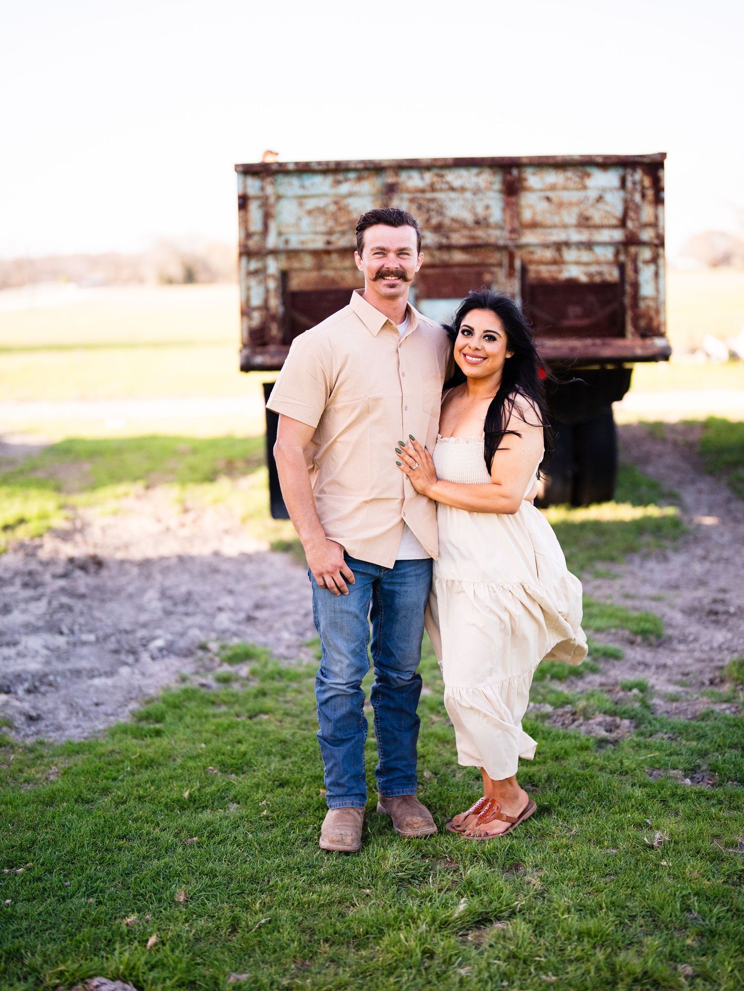 A couple poses together by a rustic wooden structure in a field during a sunny engagement photoshoot.