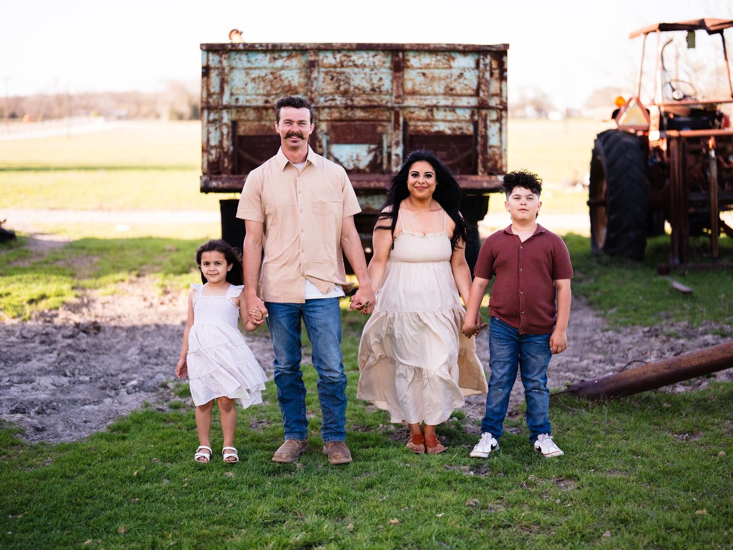 Family of four standing together in front of rustic farm equipment during sunset photoshoot.