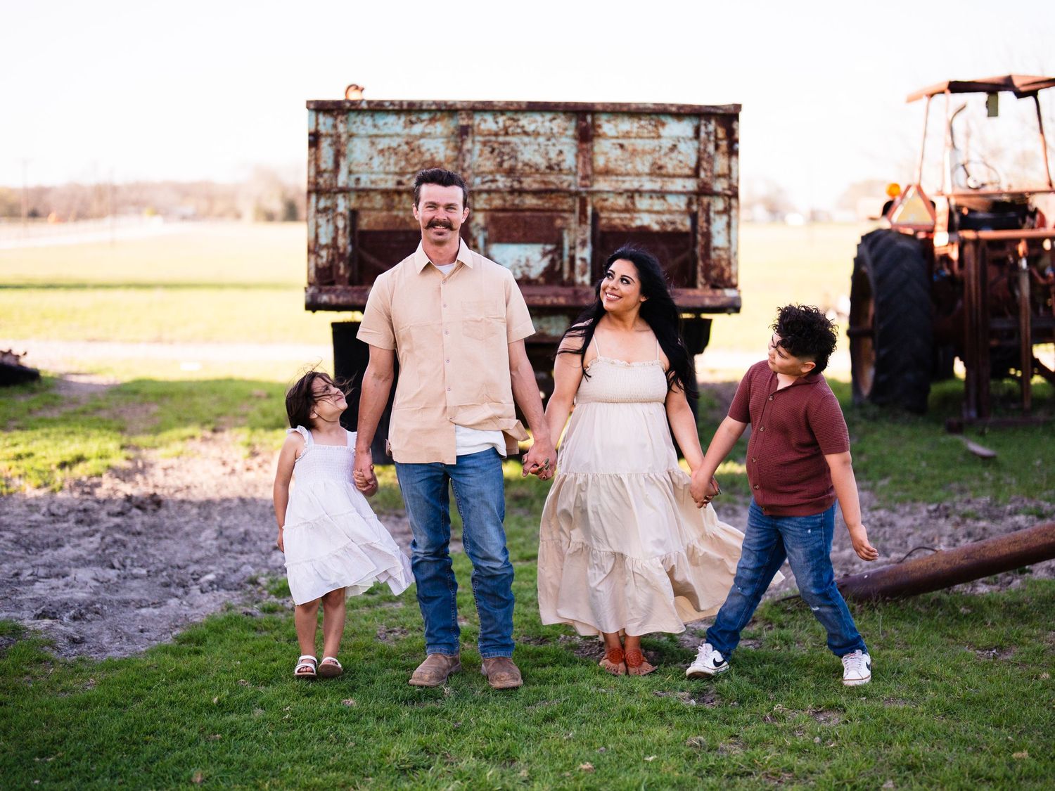 A family walks together in a rural setting with vintage farm equipment and a rustic truck in the background.