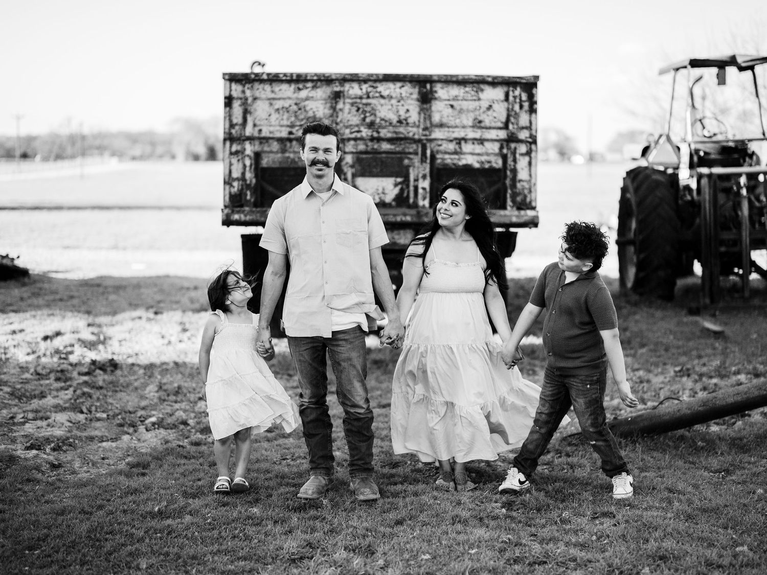 A family walks together in front of an old rustic truck in a black and white countryside setting.