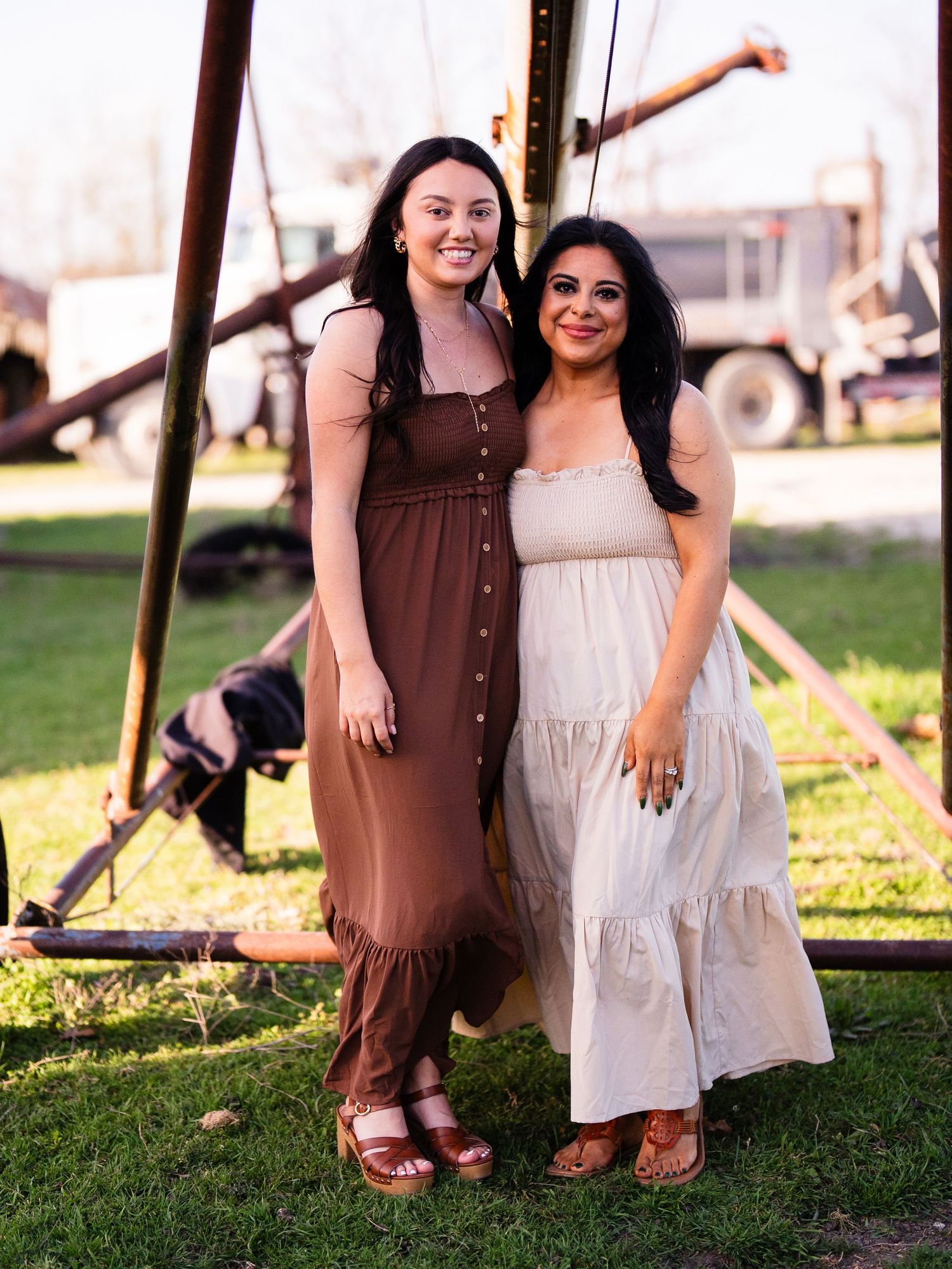 Two friends in flowing maxi dresses smile together on a grassy lawn during sunset.
