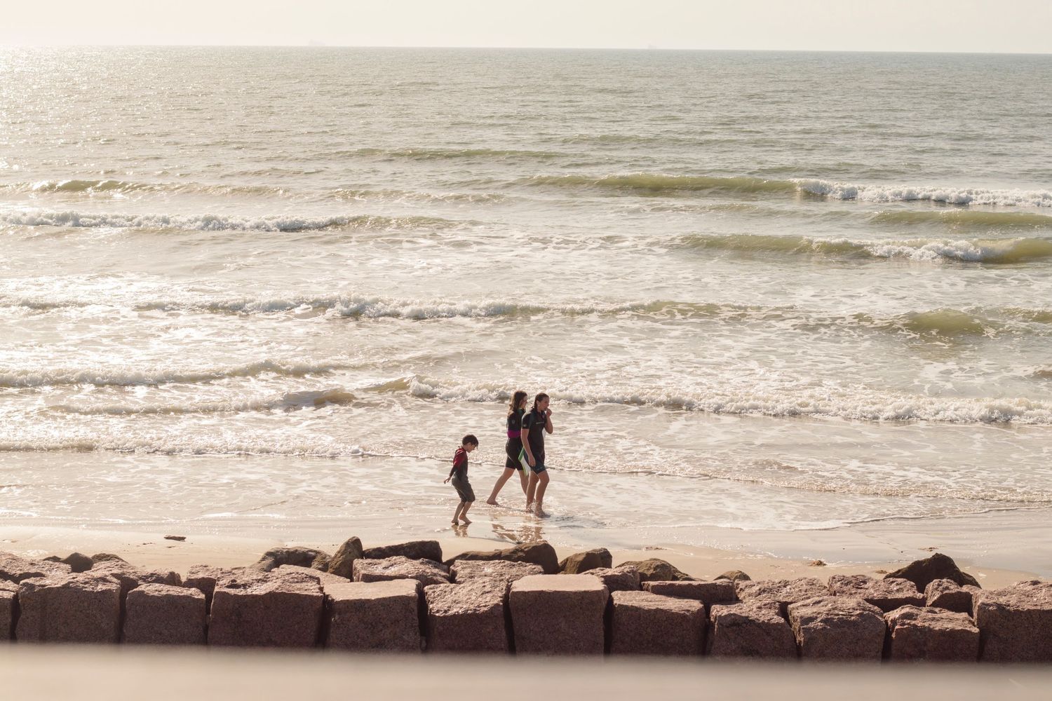 Early morning in Surfside Texas, three kids in wetsuits walk along an empty beach with rocks and crashing waves