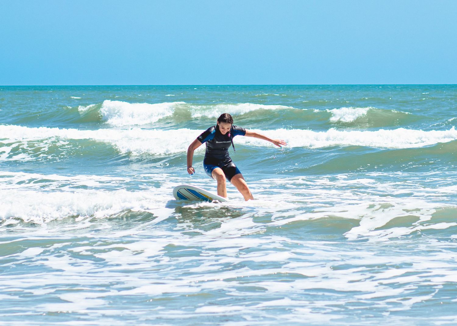 A beginner surfer rides a wave in a black wetsuit on turquoise ocean waters with white foam under bright blue skies. Surfside, Texas